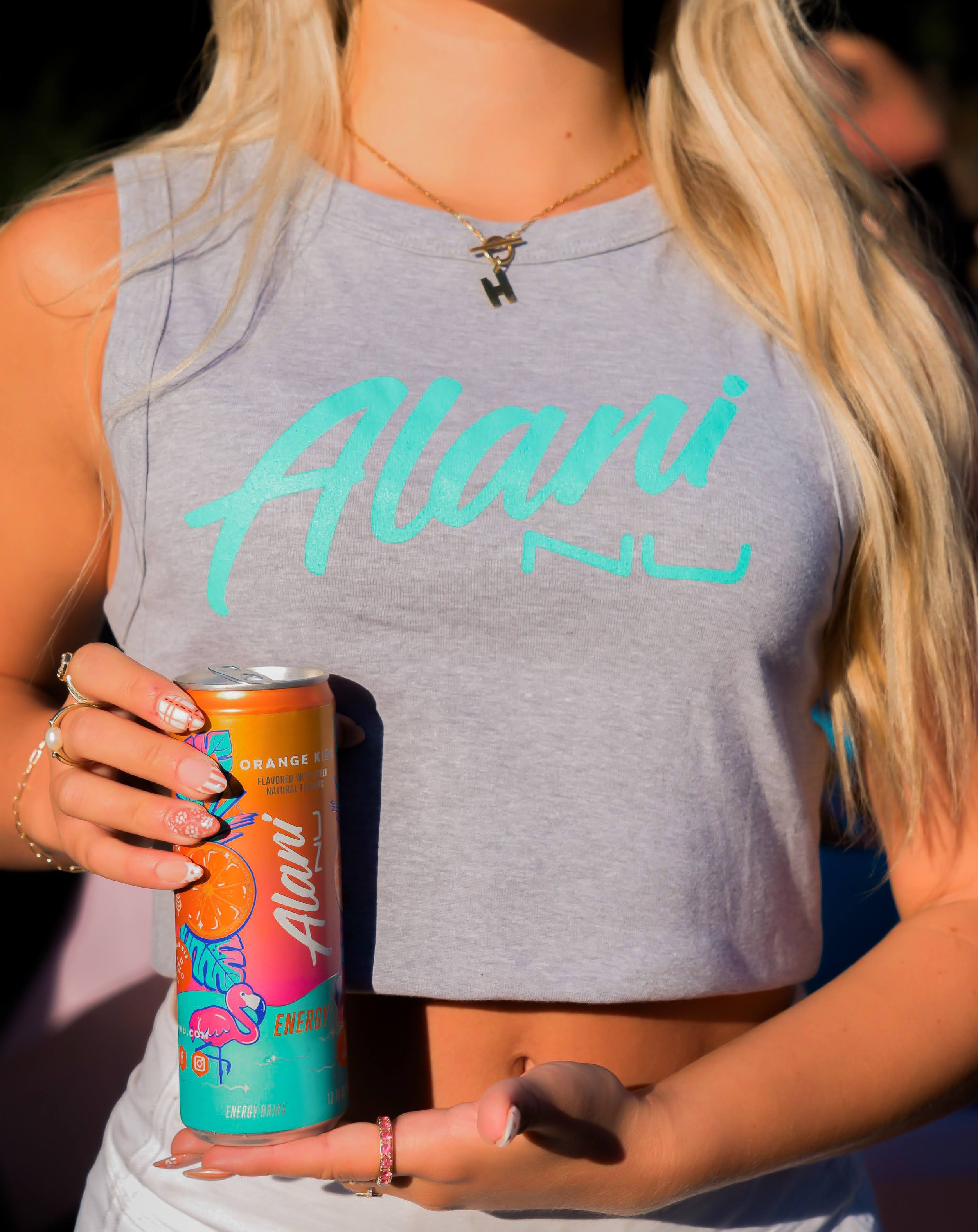 A woman wearing a gray sleeveless crop top with the name 'Alani' written on it, holding a can of Alani energy drink with a colorful flamingo and orange slice design, wearing a gold necklace with a 'H' pendant and a small bird charm, and decorated wit