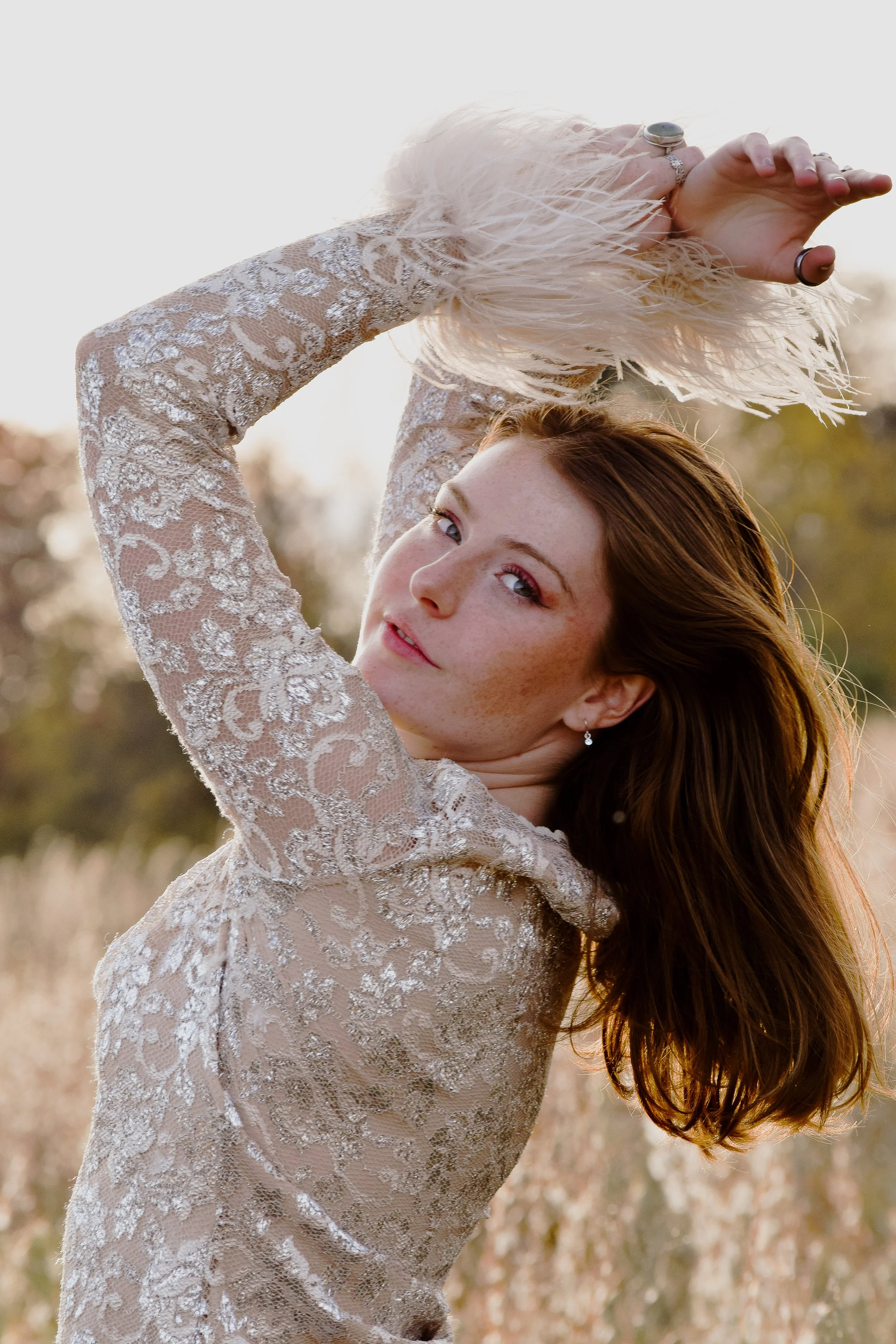 A woman with red hair and freckles, wearing a sheer lace dress, holds a feathered fan above her head outdoors during golden hour.