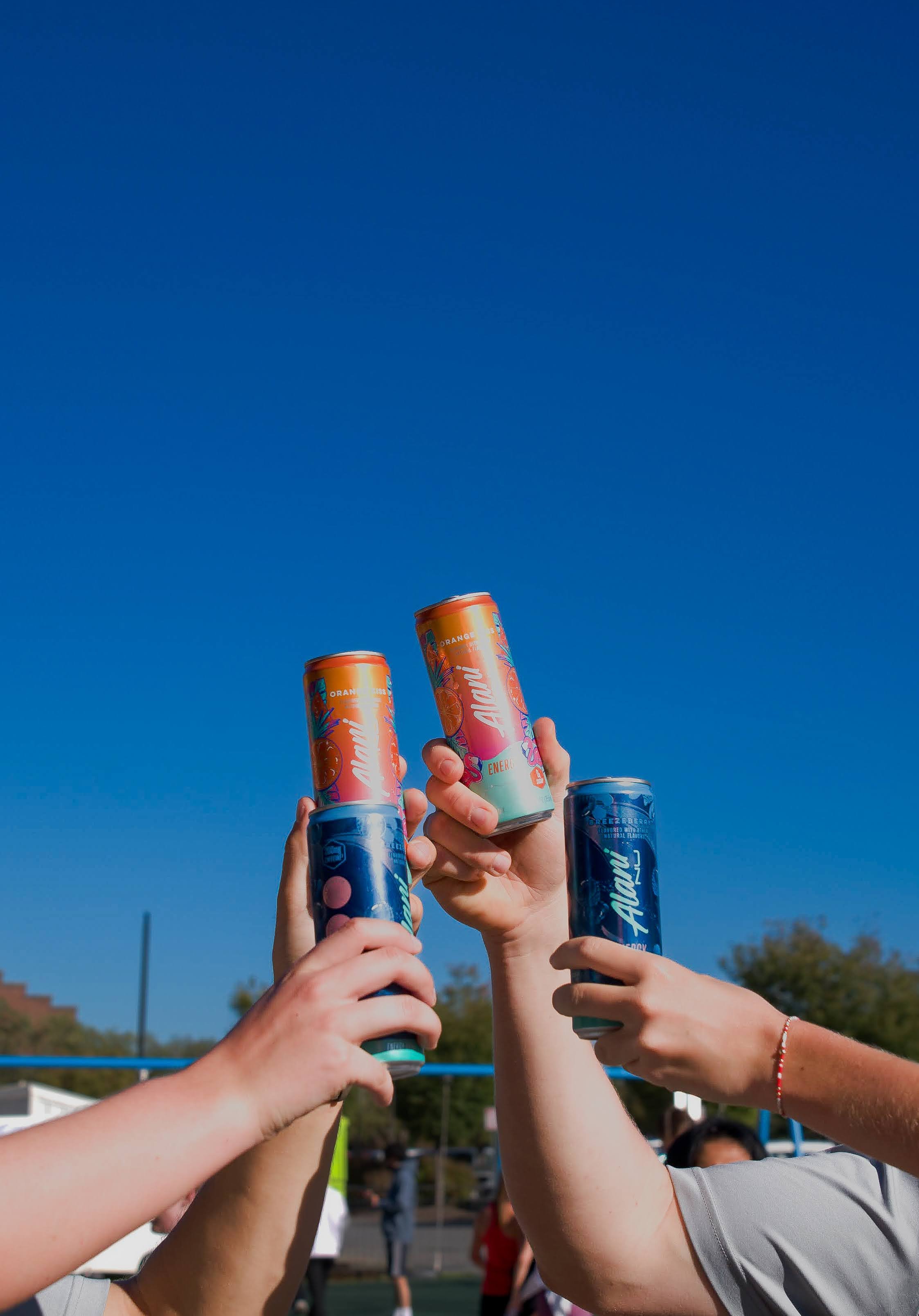 Several hands holding up cans of Alani energy drinks with a clear blue sky in the background.