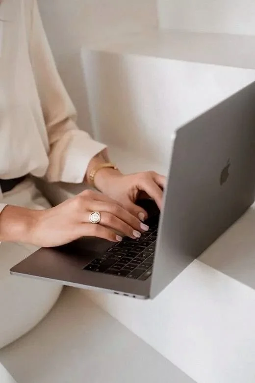 Person typing on a silver MacBook laptop, wearing a ring and a gold bracelet, sitting at a white surface.