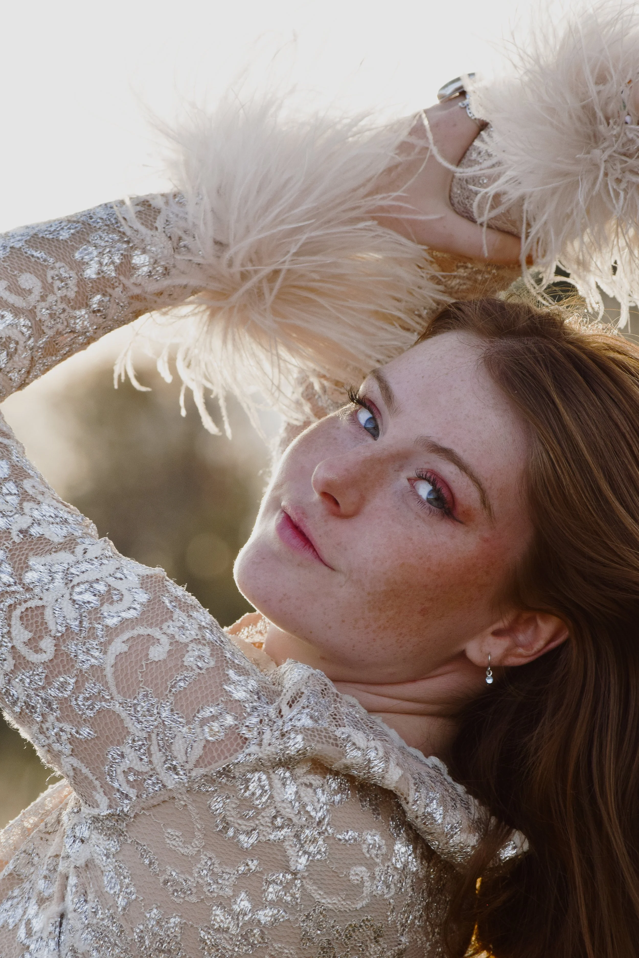 A woman with red hair and freckles wearing a lace dress with feathers, looking at the camera outdoors during sunset.