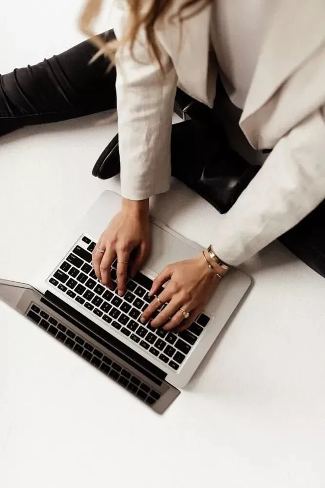 Person in a beige blazer typing on a silver laptop on a white table, with another laptop reflected below.