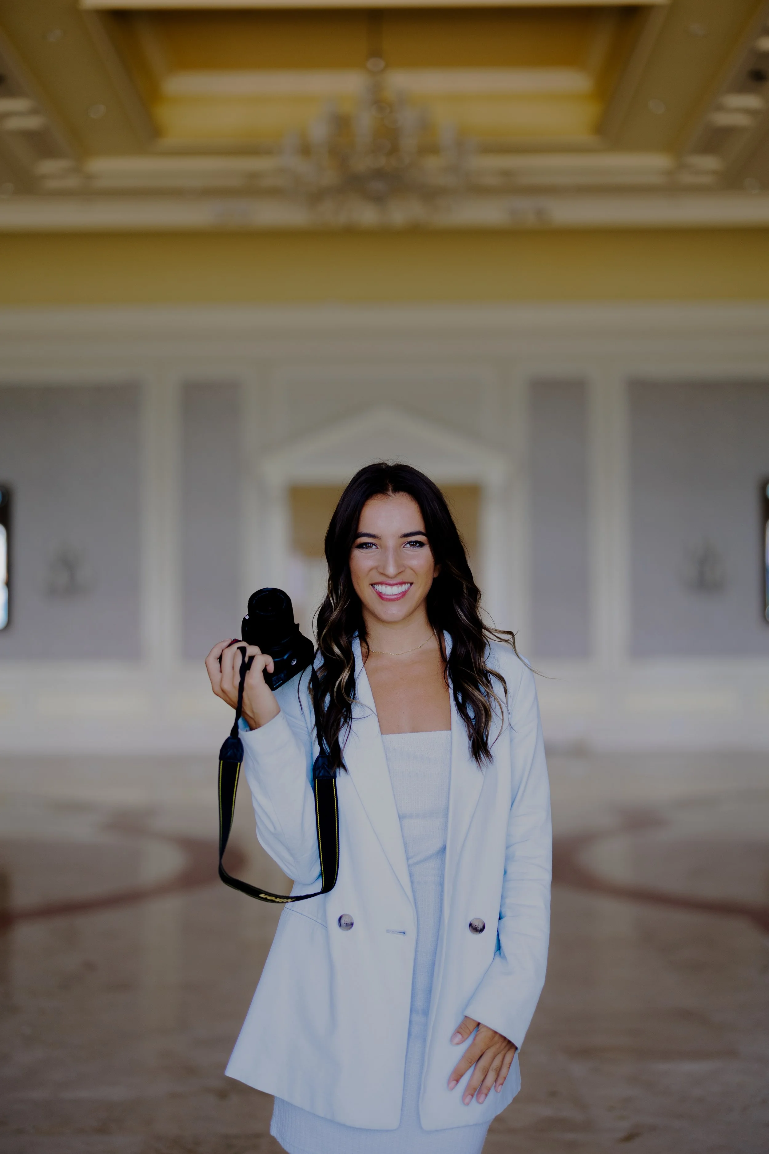 A woman with long dark hair is smiling and holding a camera, standing in an elegant, well-lit indoor setting with high ceilings and decorative details.