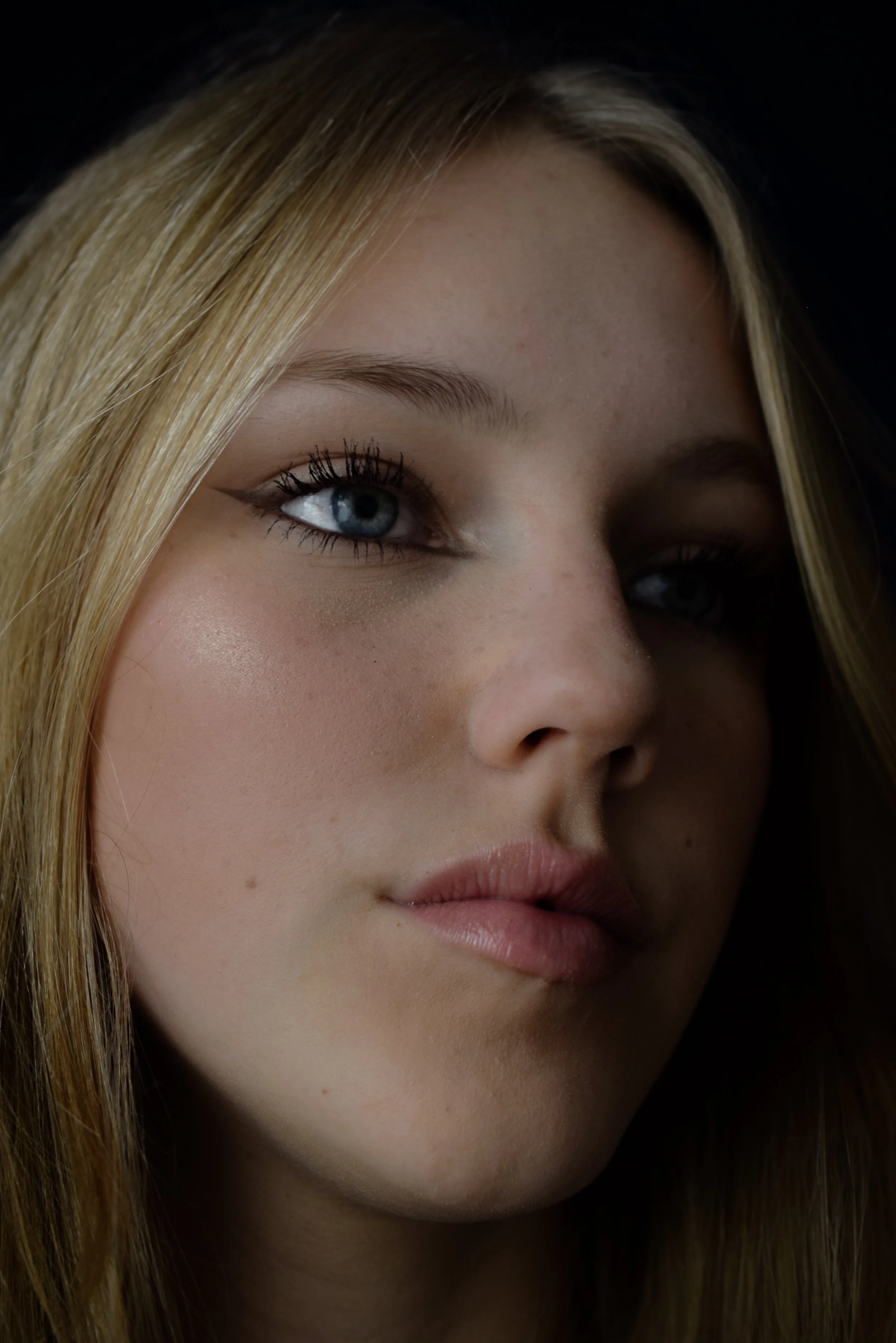 Close-up portrait of a young woman with blonde hair and blue eyes, makeup including eyeliner and lipstick, looking softly at the camera.