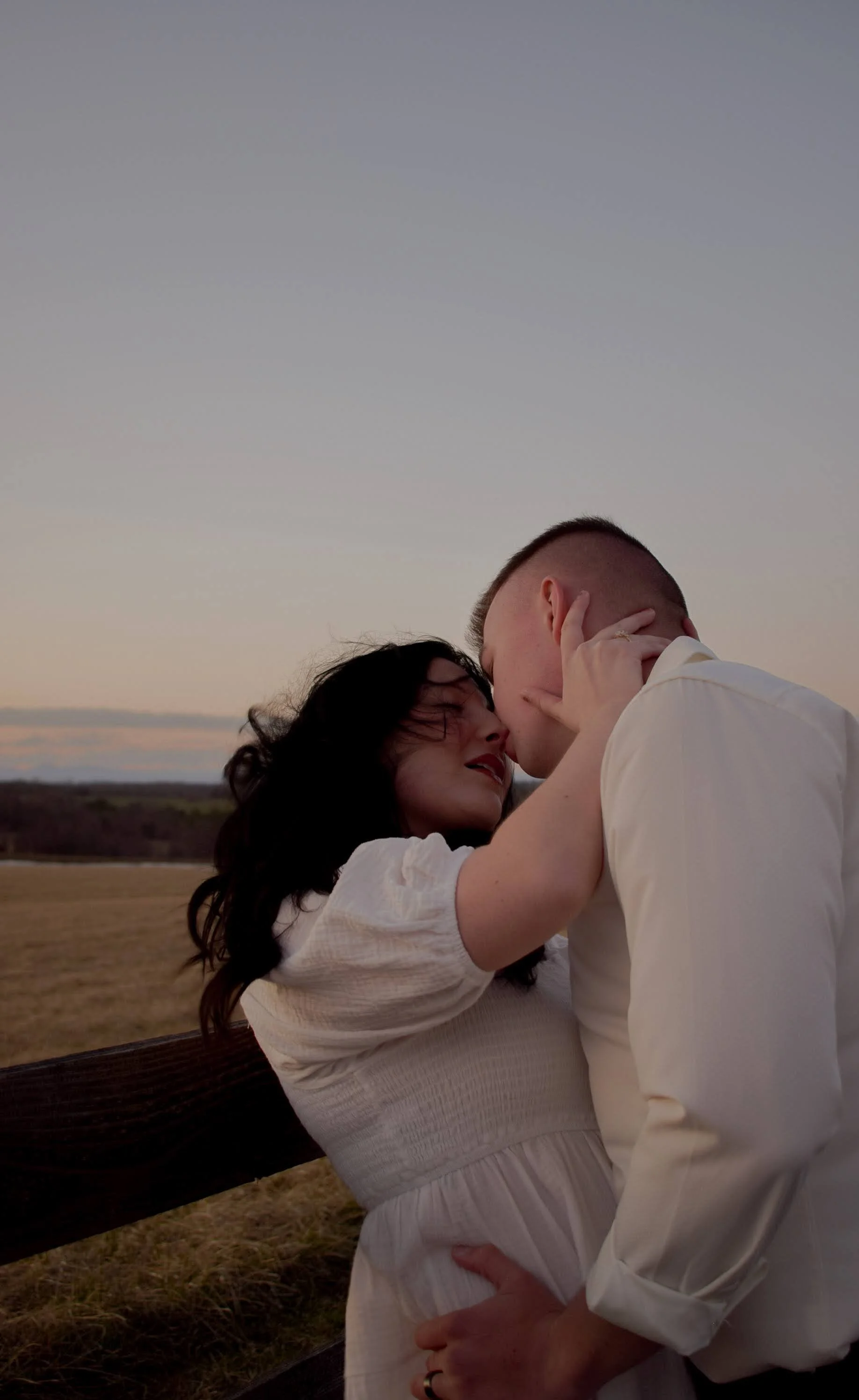 A couple sharing a romantic moment outdoors at sunset, with the woman holding the man's face and leaning in for a kiss.