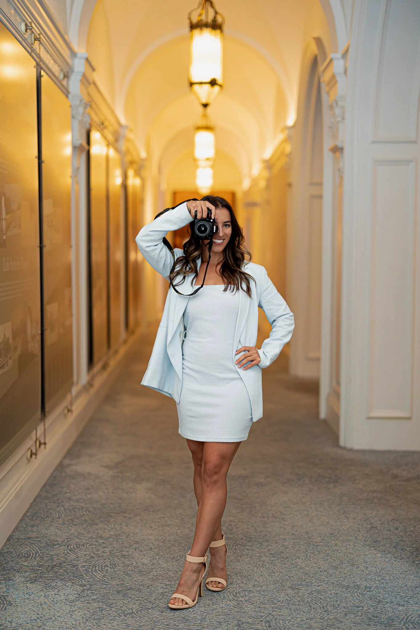 A woman with long, wavy brown hair smiling while taking a photo with a camera in a well-lit, elegant hallway with arched ceilings and ornate lighting fixtures.