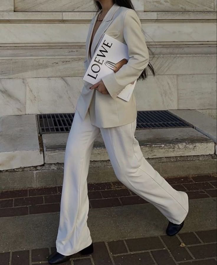 A person walking outdoors, wearing a beige suit with wide-legged pants, holding a Loewe shopping bag and a folder, with their face not visible.
