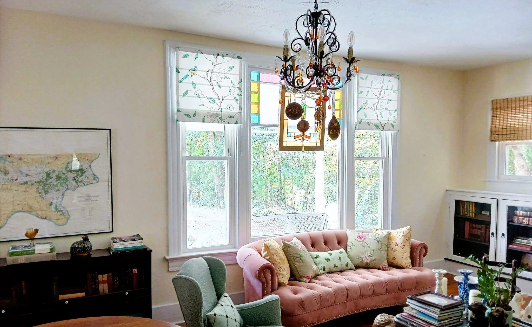 Living room with large window, pink velvet sofa with floral and patterned pillows, black chandelier, map on wall, bookshelves, and decorative items.