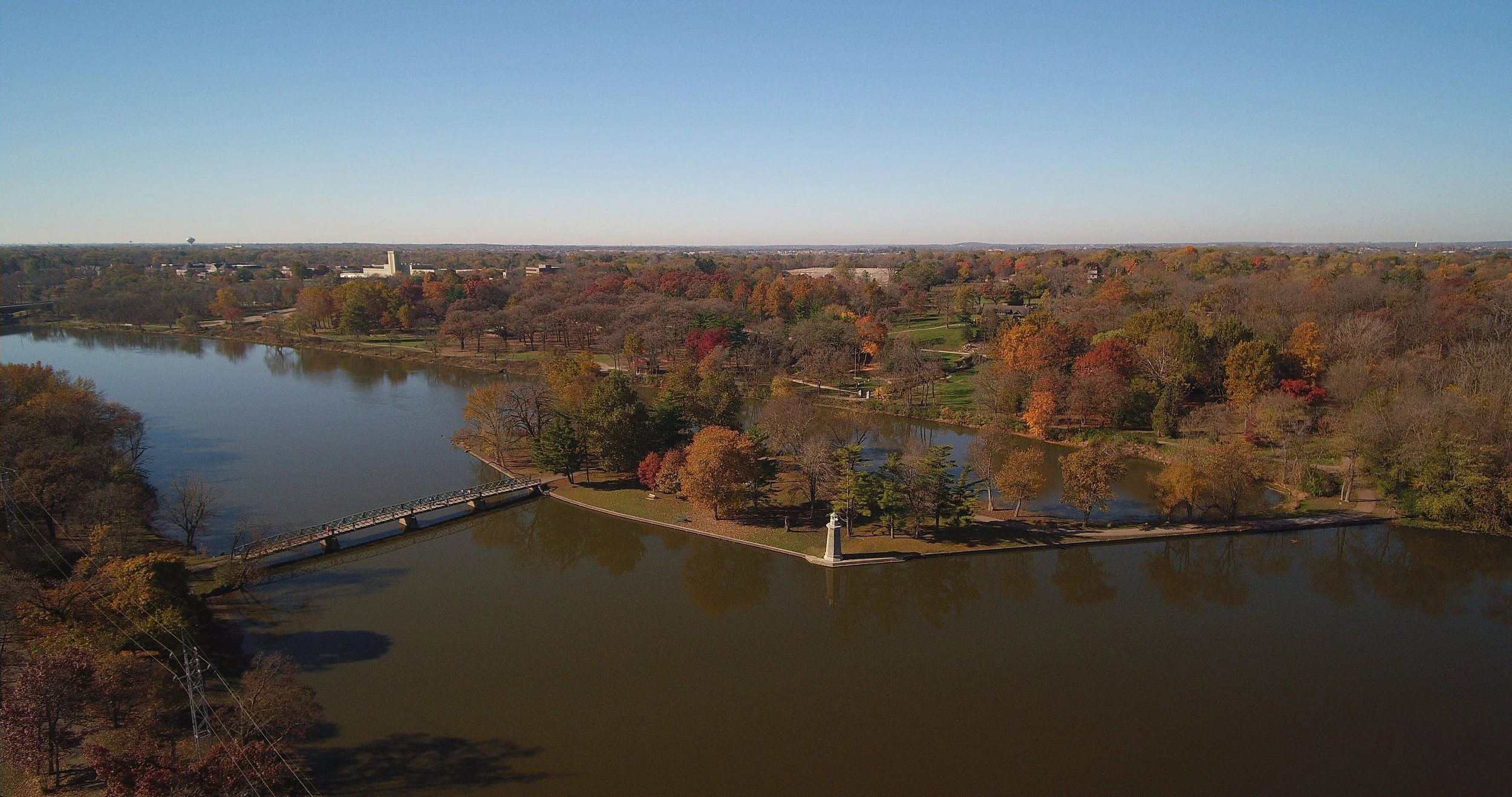 Aerial view of a park with a large lake, trees with autumn foliage, a small lighthouse, and a bridge crossing the lake, with a city skyline in the background under a clear blue sky.
