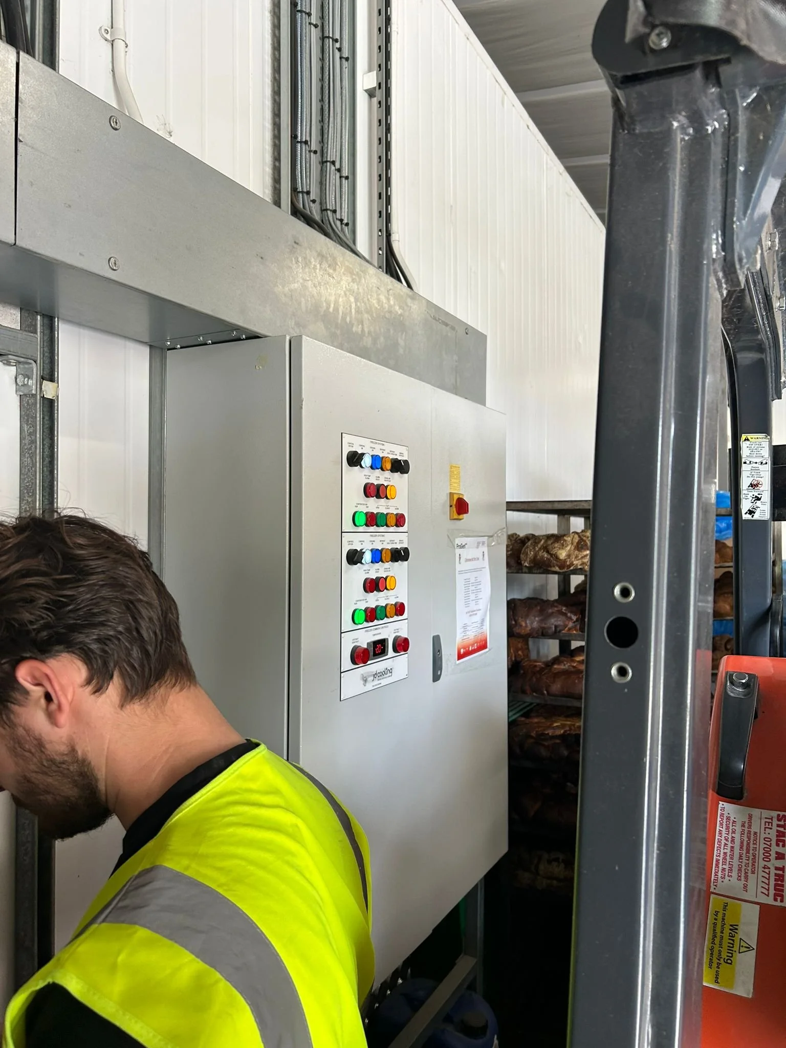 A worker in a high-visibility vest working near an electrical control panel inside a storage or industrial facility.