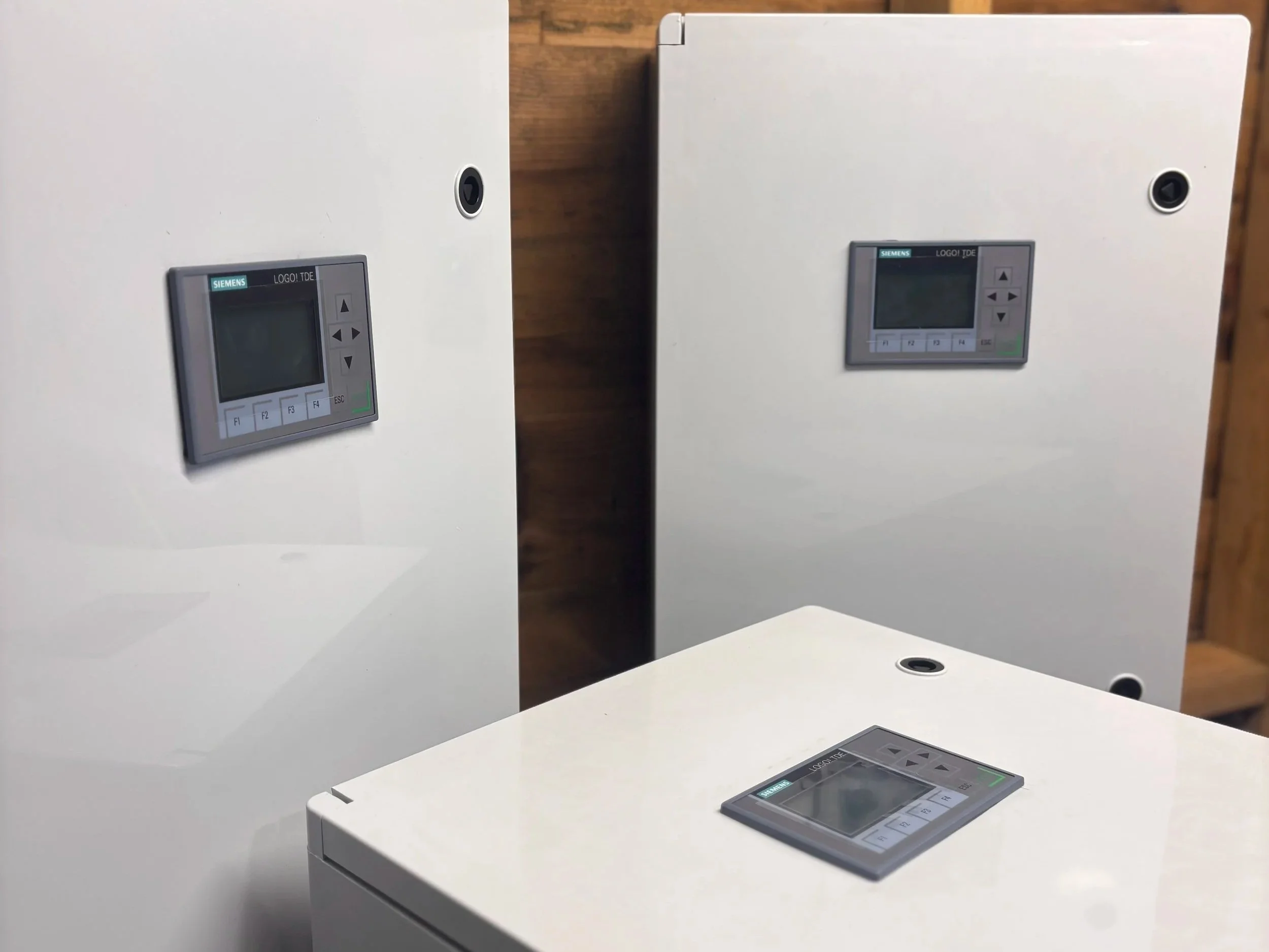 Three white electrical control panels with Siemens digital displays mounted on a wooden wall and a white surface.
