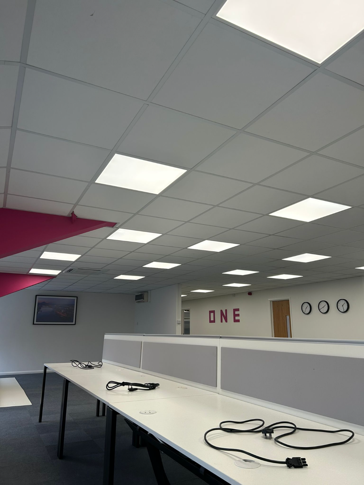 Empty office space with a ceiling of white tiles, fluorescent lighting, a long white table with black cords, a wall with large pink letters spelling "ONE," and three clocks showing different times.