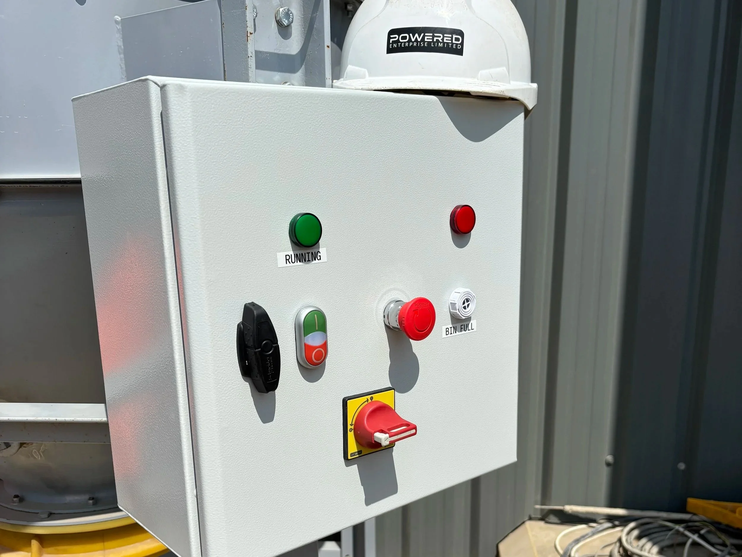 An industrial control panel with buttons and switches, including a green start button labeled "RUNNING," a red stop button, and a red emergency stop switch, with a white hard hat labeled "POWERED ENTERPRISE LIMITED" resting on top.