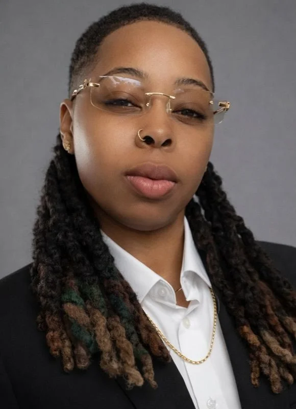 A woman with long dreadlocks, wearing glasses, a nose ring, earrings, a white collared shirt, a black blazer, and a gold chain, posing against a gray background.