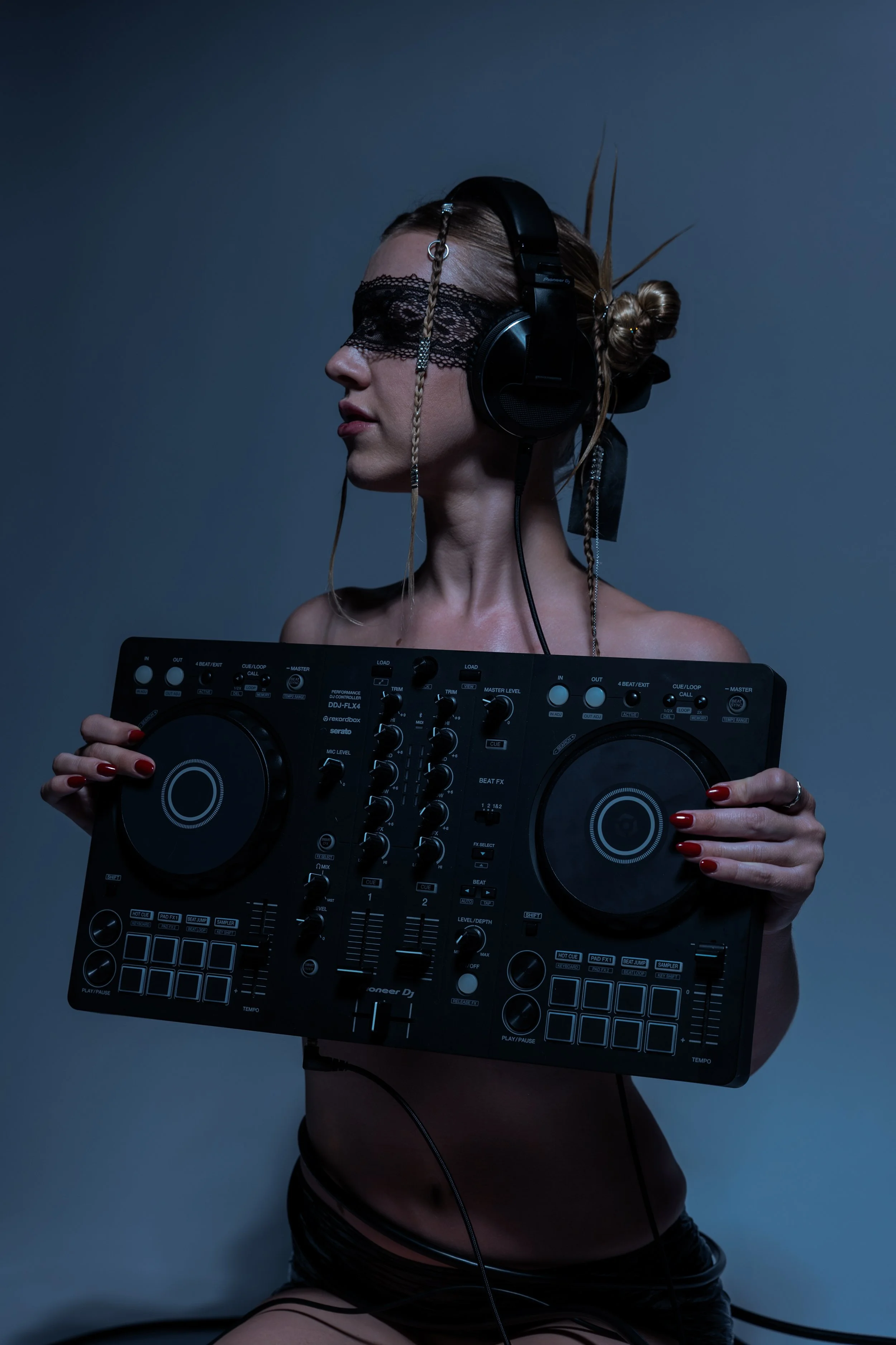 Claire De Lune wearing headphones and a lace eye mask, holding DJ equipment, with her hair styled in buns and braids, against a dark background.
