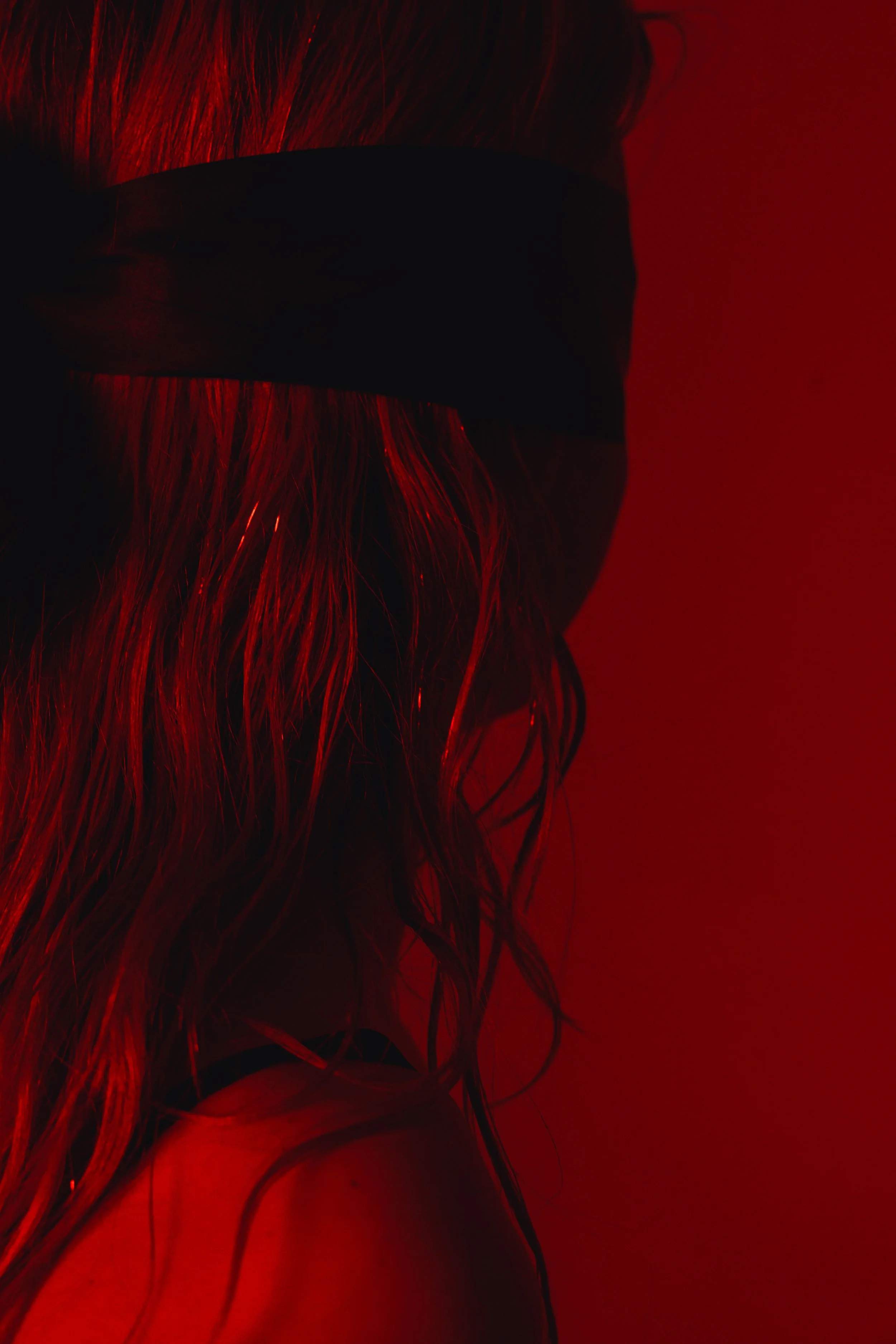 A close-up profile of Claire De Lune with long, wet, hair wearing a black mask and black strap, against a red background.