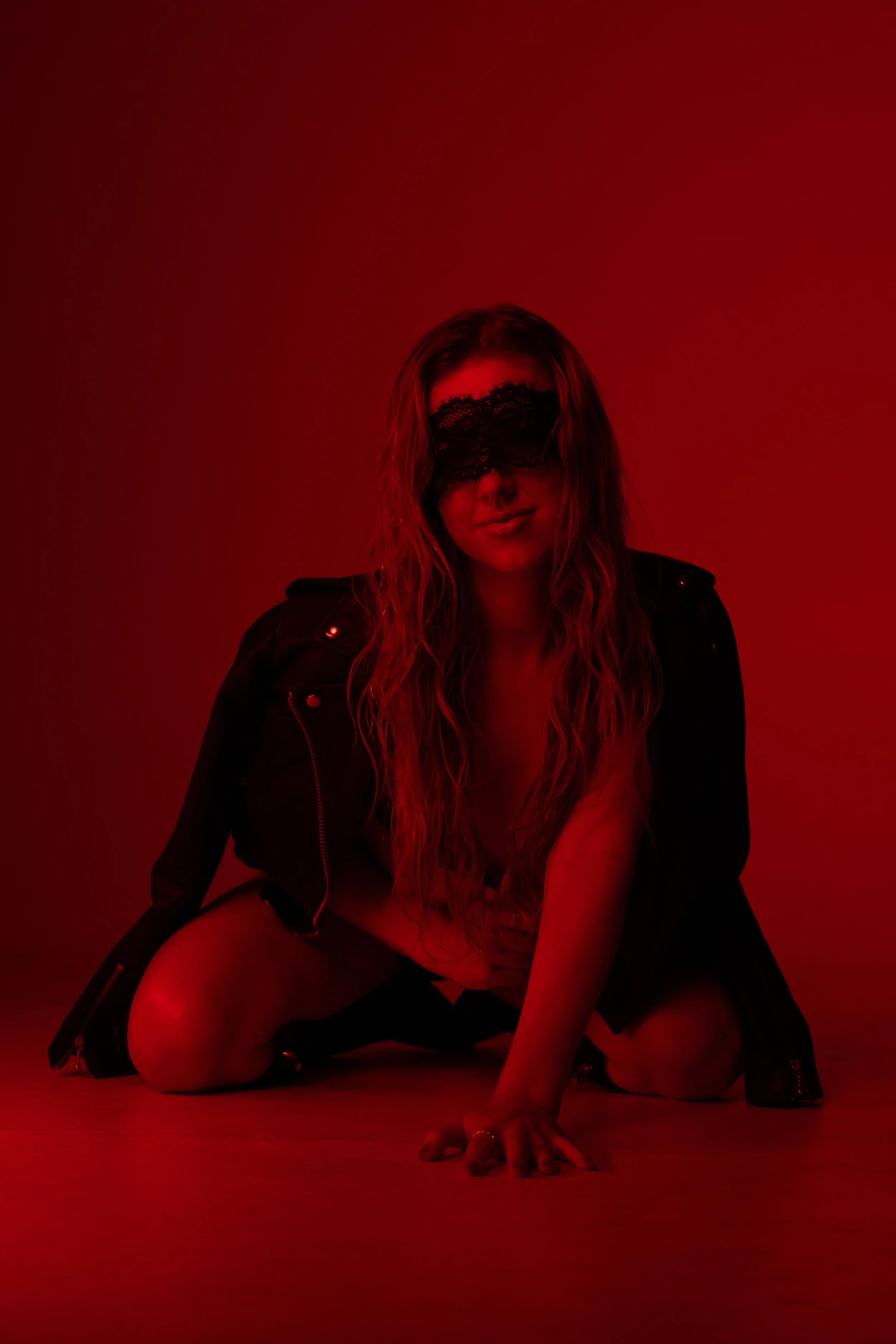 Claire De Lune with long, wavy hair dons a black lace eye mask and a black leather jacket, kneeling on the floor in a red-lit setting with a dark background.