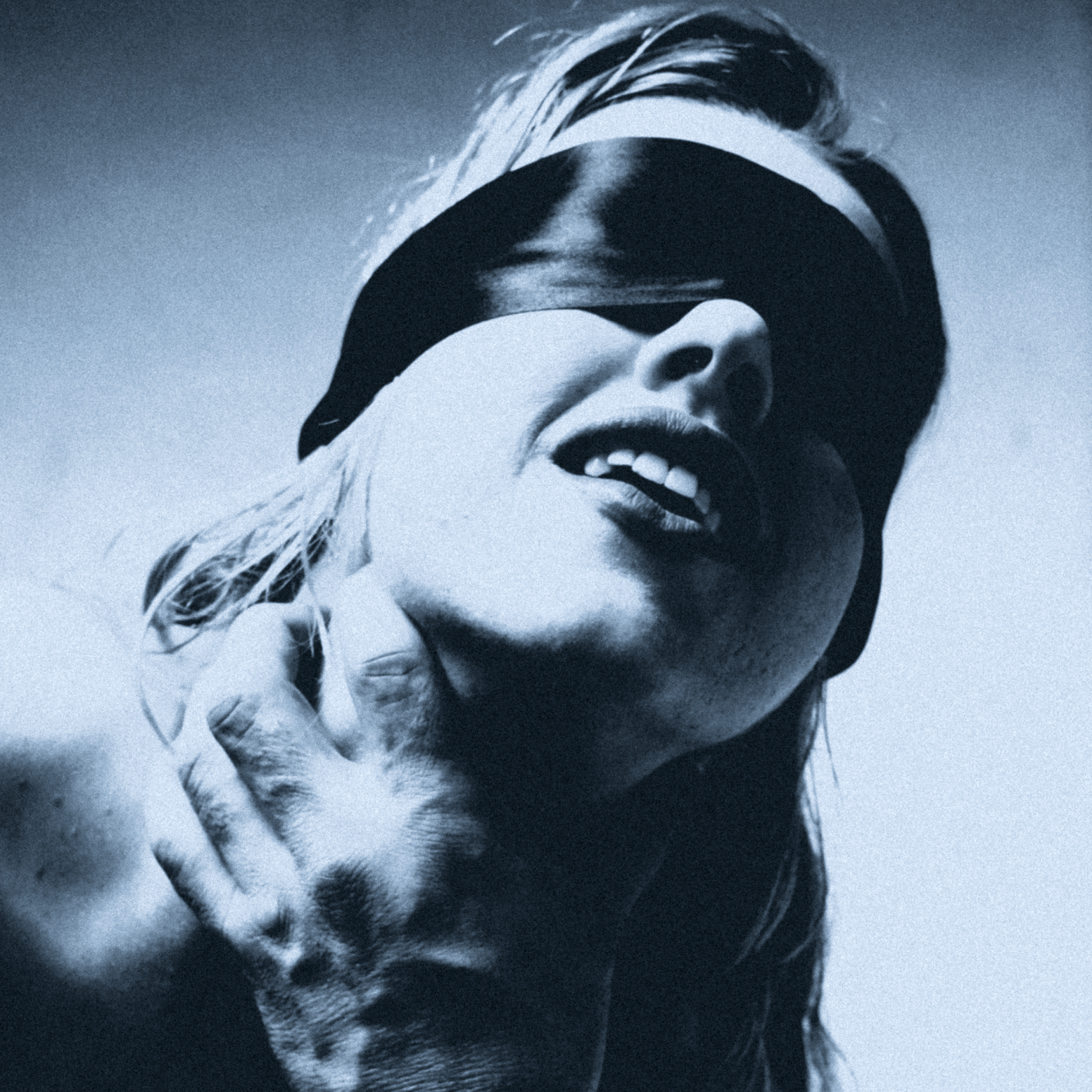 A black and white photo of Claire De Lune with tousled hair wearing a blindfold, touching her neck with her hand, and smiling with her mouth slightly open.