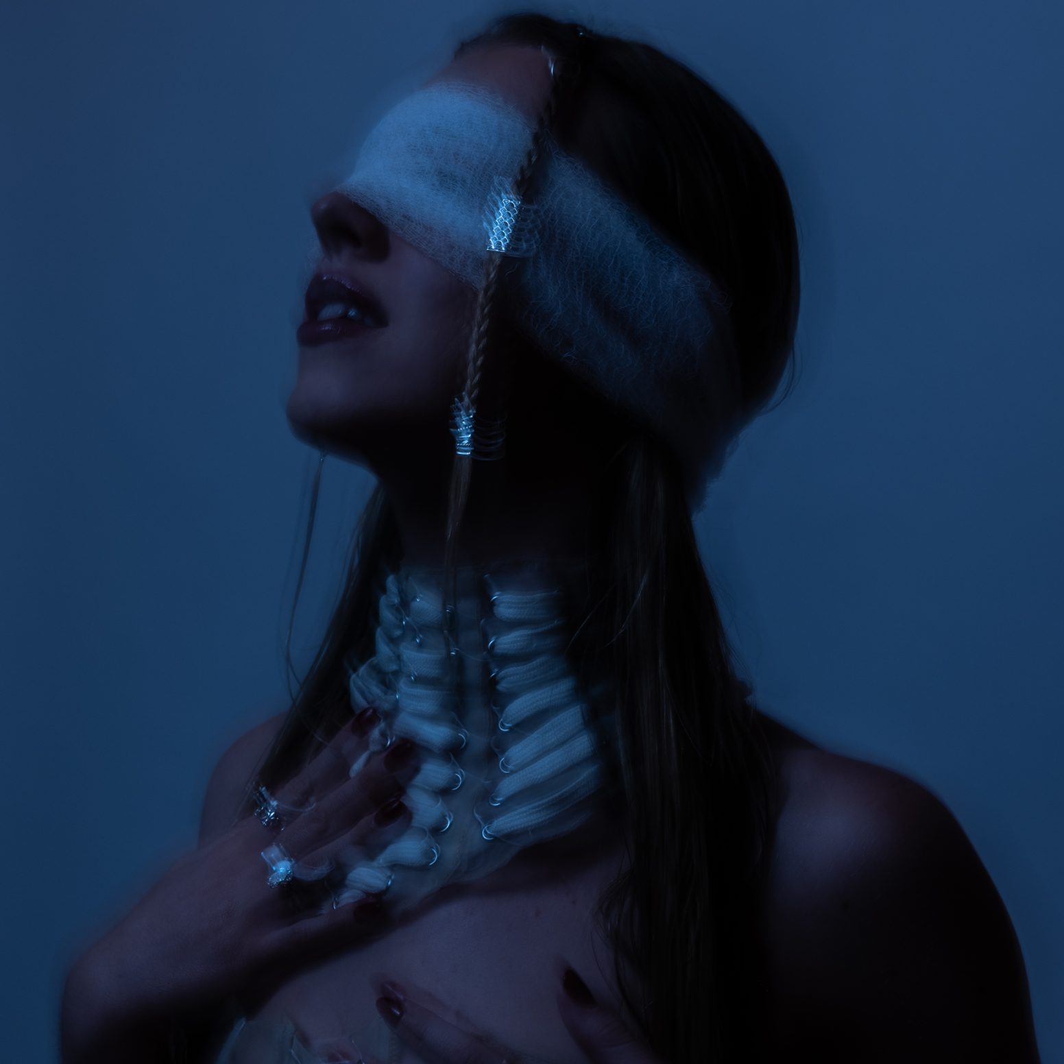Claire De Lune with a bandaged face and skeletal hand necklace, touching their chest, with long dark hair and dark lipstick, against a dark background.