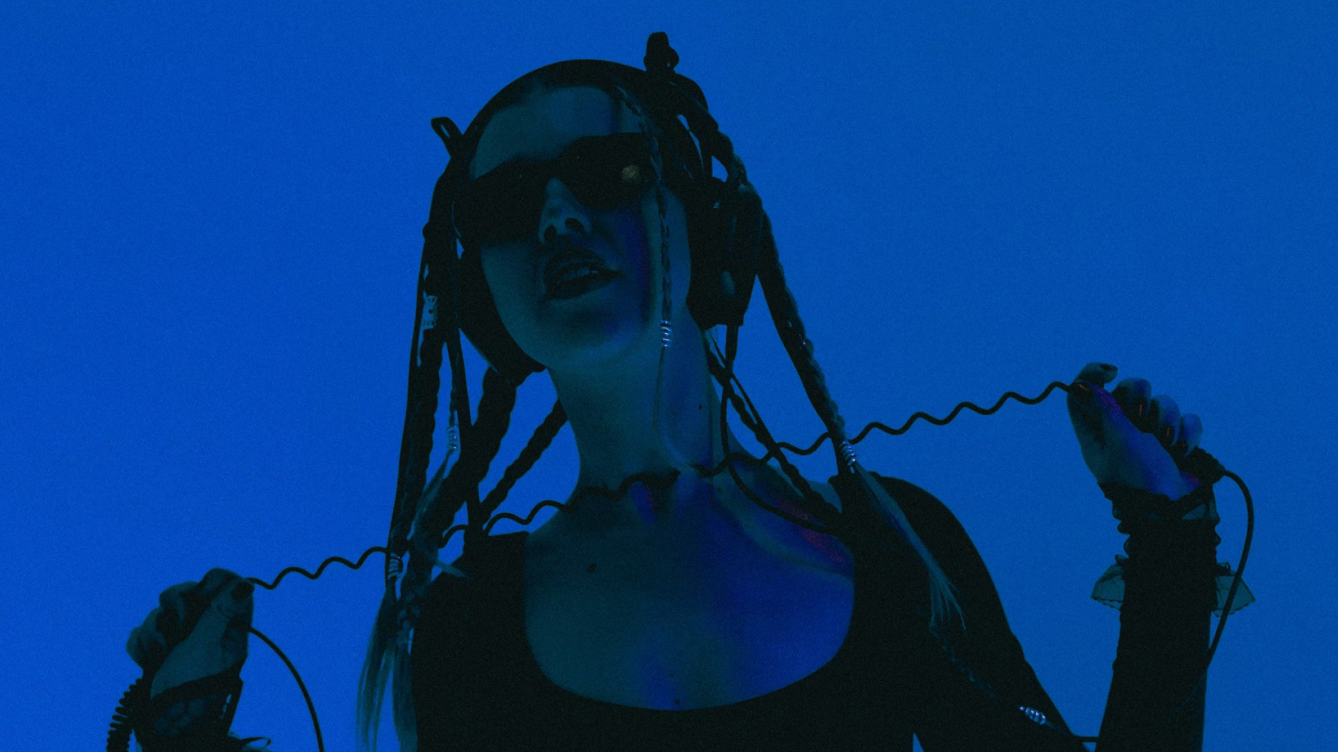 A woman wearing sunglasses and black clothing is holding a vintage telephone cord in both hands, with a blue background.
