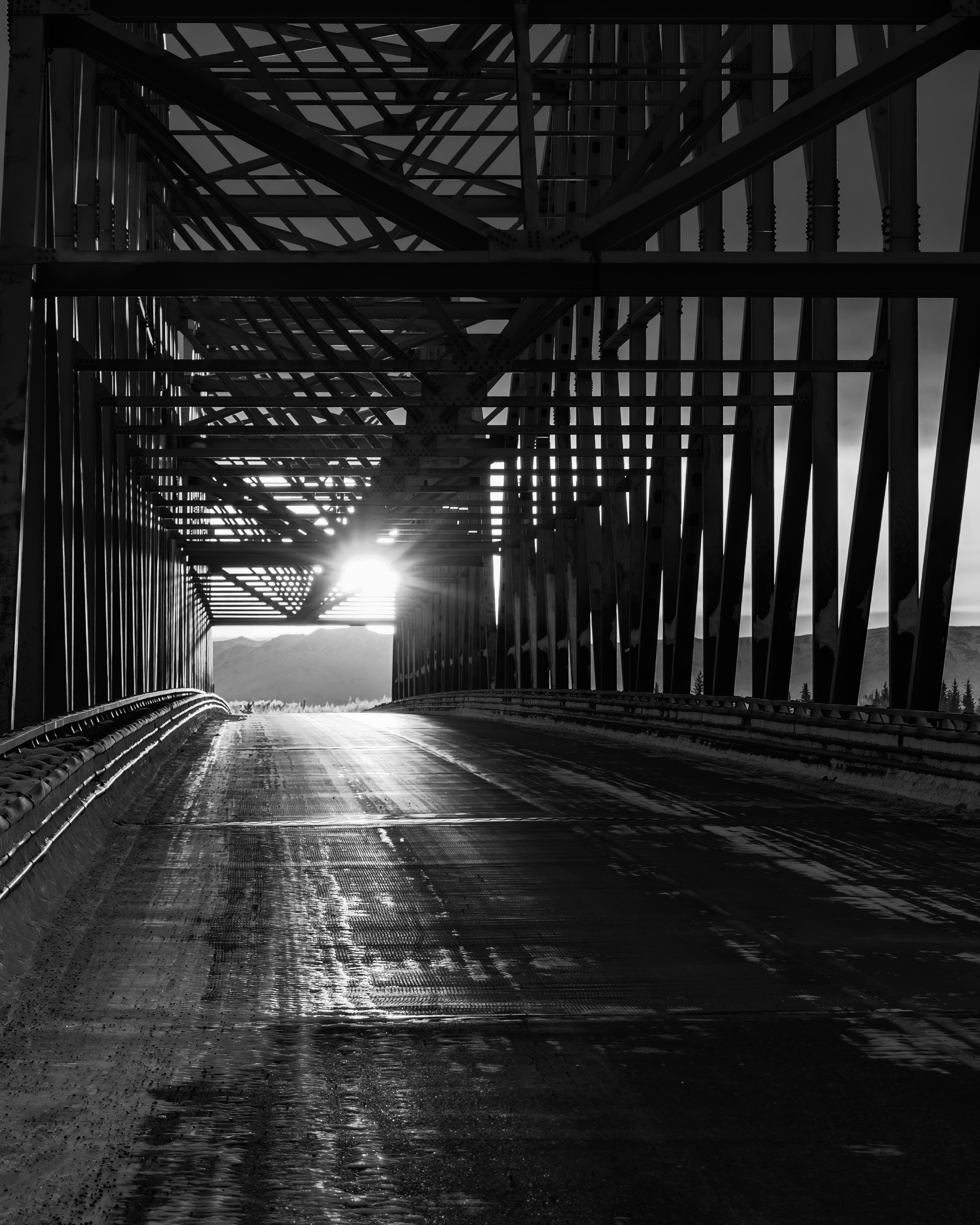 Under a bridge with steel beams, a road curves into the distance, sunlight shining through the gap in the structure.