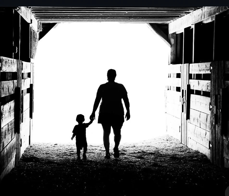 Black and white photograph of an adult and a child holding hands, walking toward light through an open doorway.