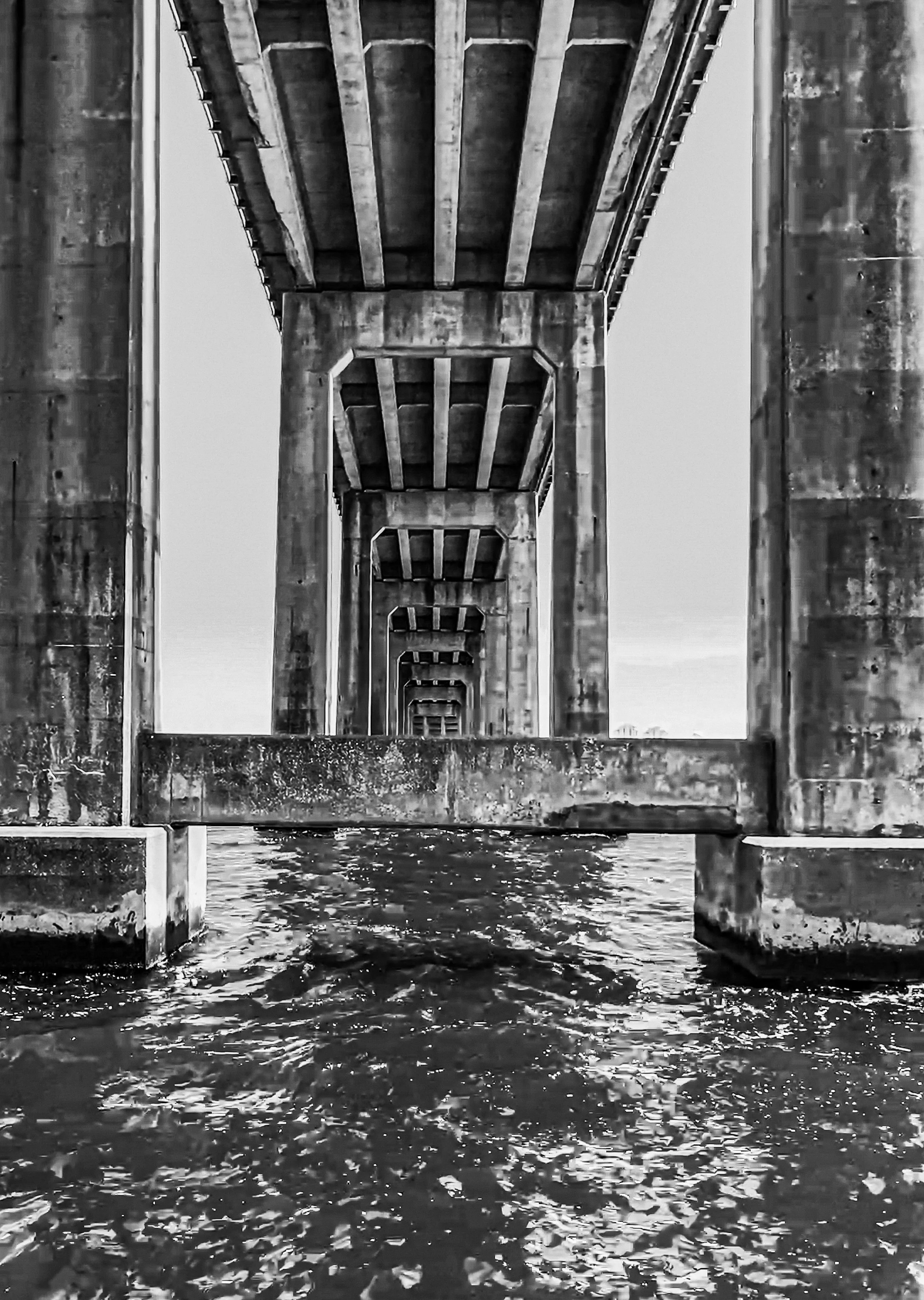 Black and white photograph taken beneath a bridge, showing repeating concrete columns and beams over water.
