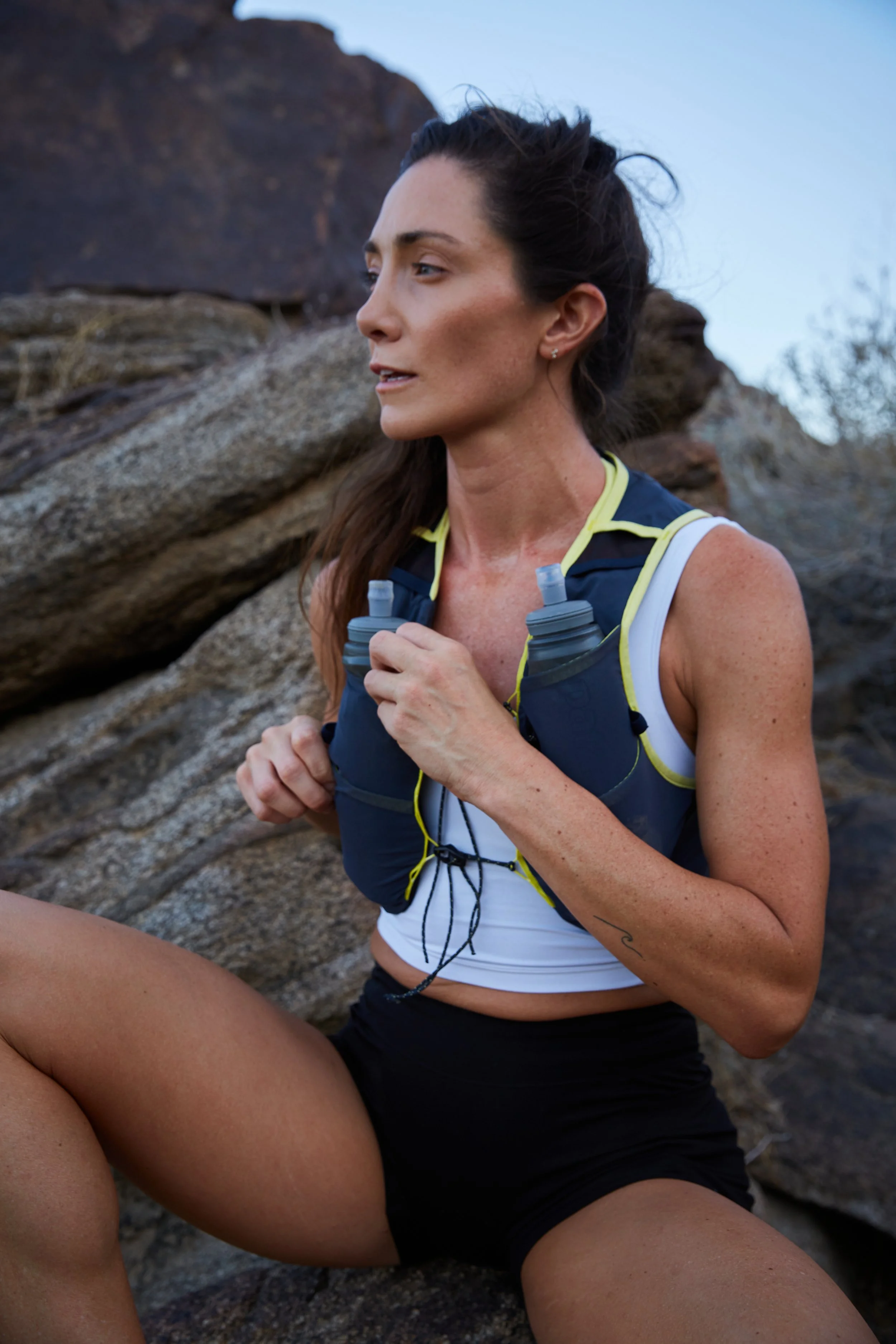 A woman with long dark hair in a ponytail, wearing a white sports bra and black shorts, sitting on rocks outdoors, holding a water bottle, and wearing a hydration vest.