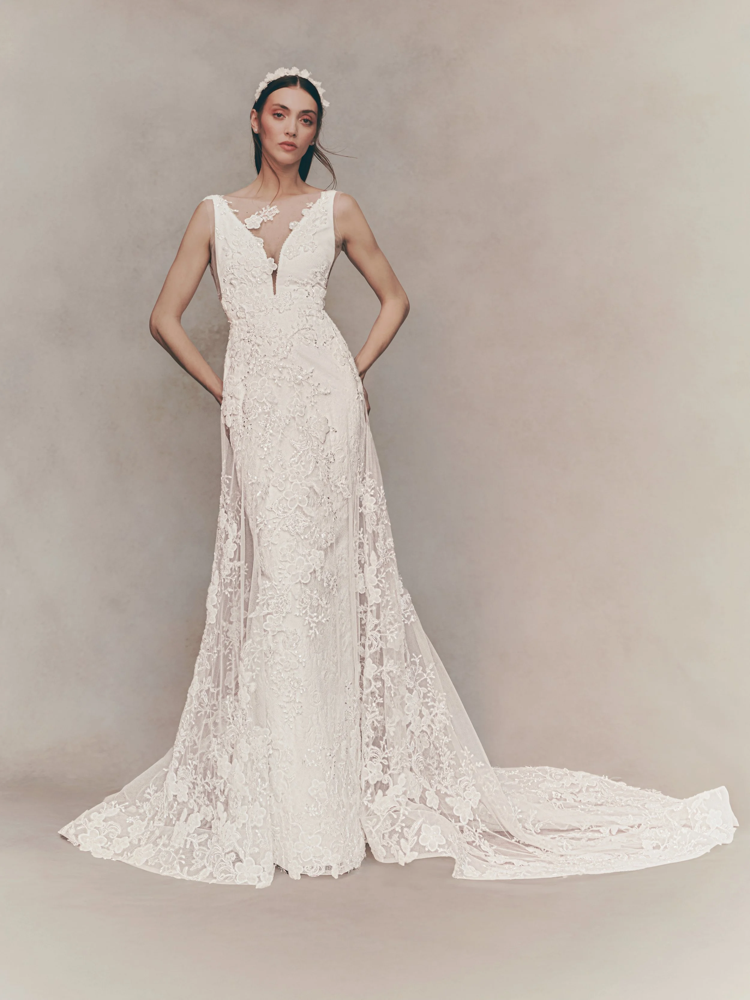 A woman in a detailed white lace wedding dress with a long train, standing against a plain background.