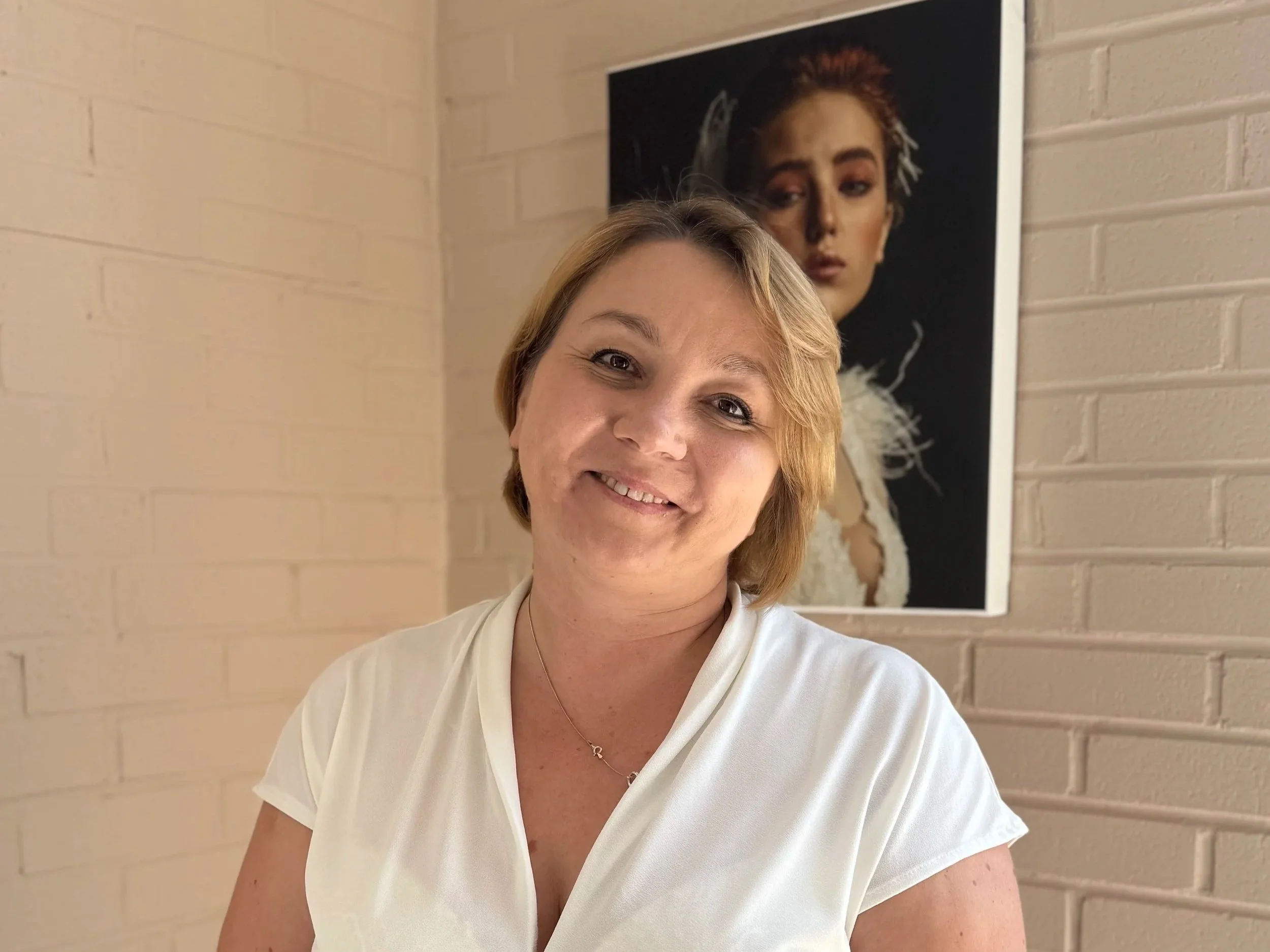 A woman with short blonde hair smiling in front of a beige brick wall, with a portrait of a woman with red hair and makeup hanging behind her.