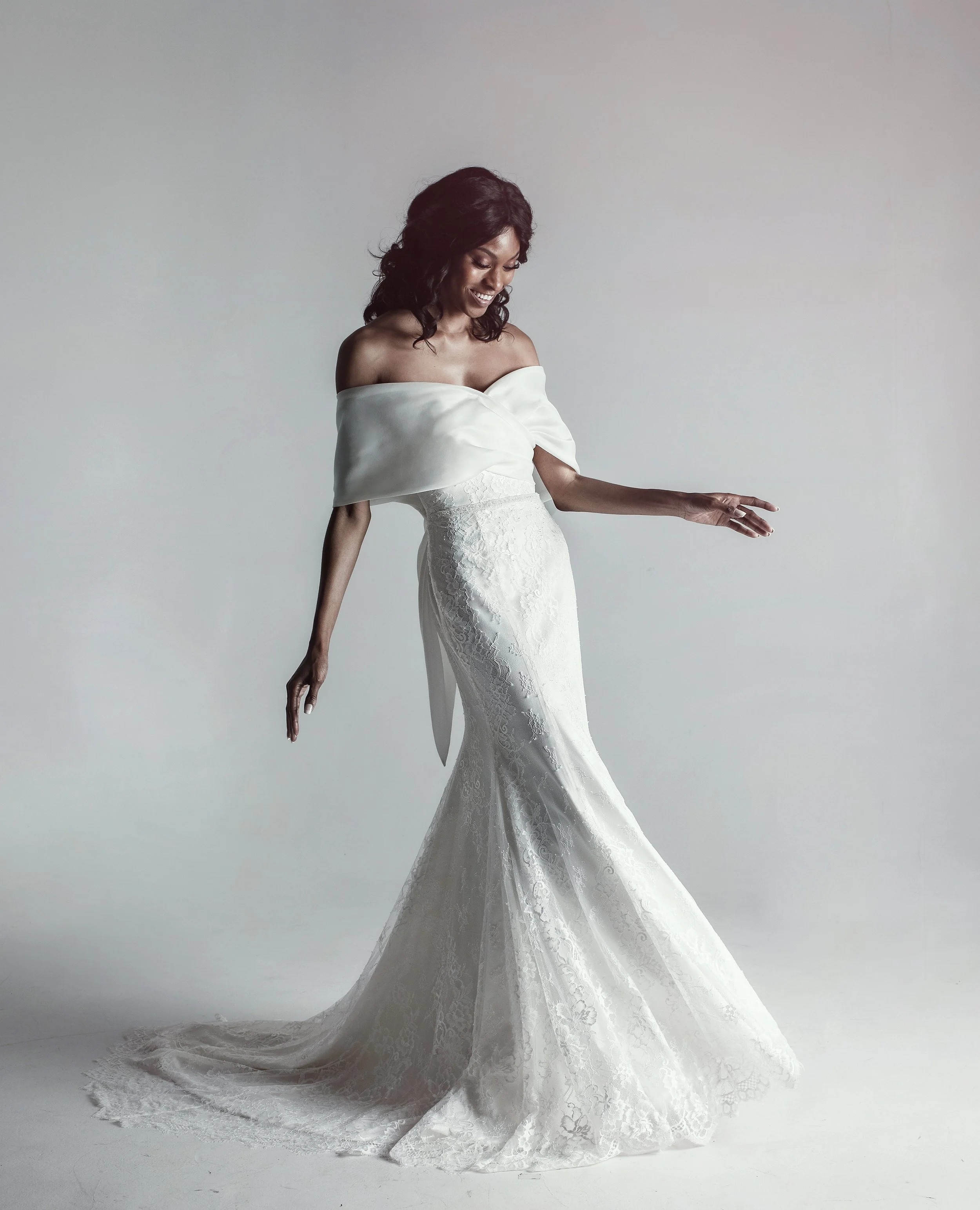 A woman in a white lace strapless wedding gown with a off-the-shoulder satin wrap, standing on a plain background, smiling and looking down.