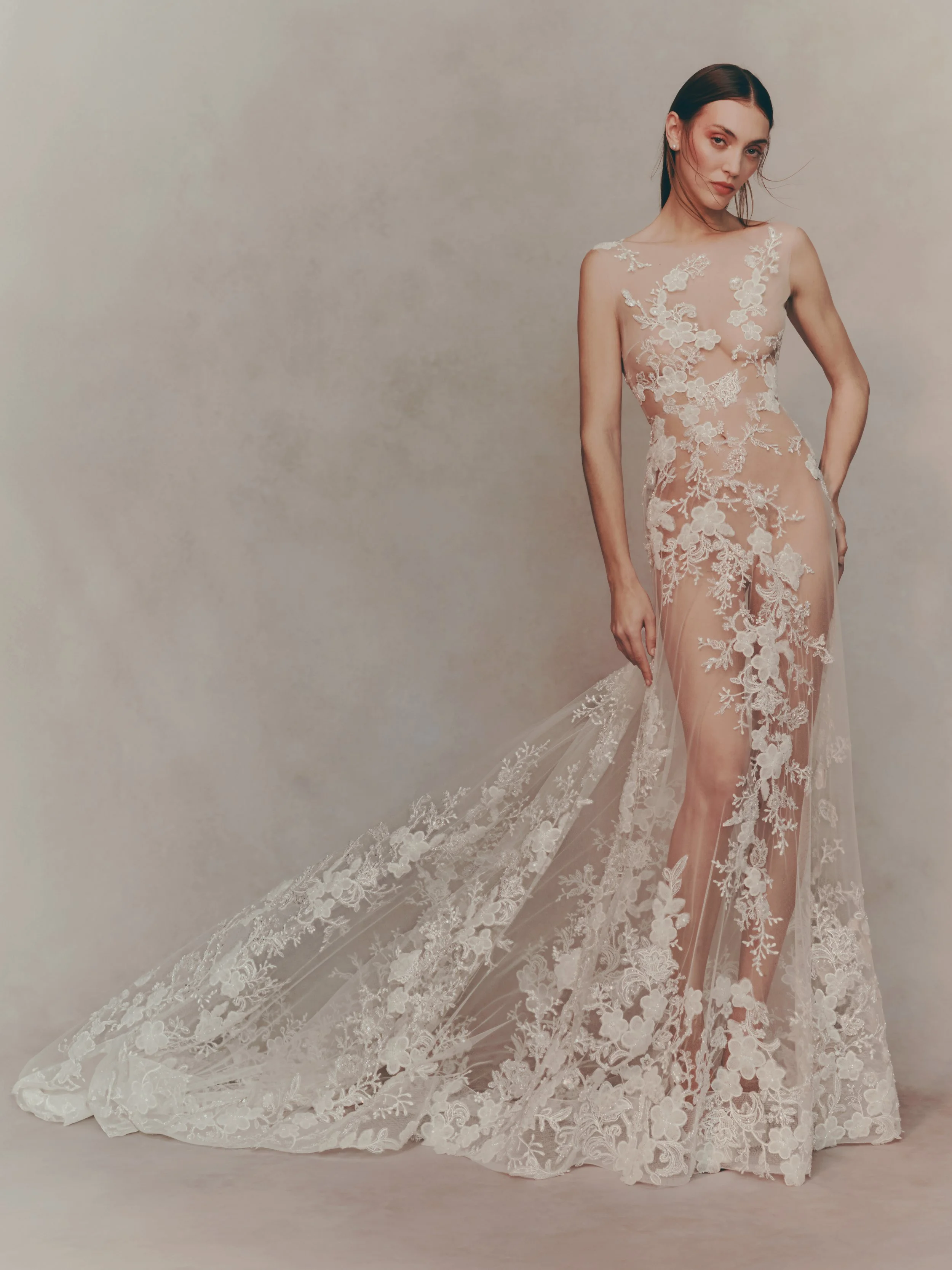 A woman in a sheer, white lace wedding dress with floral embroidery, standing against a beige background, holding the fabric train of her dress.