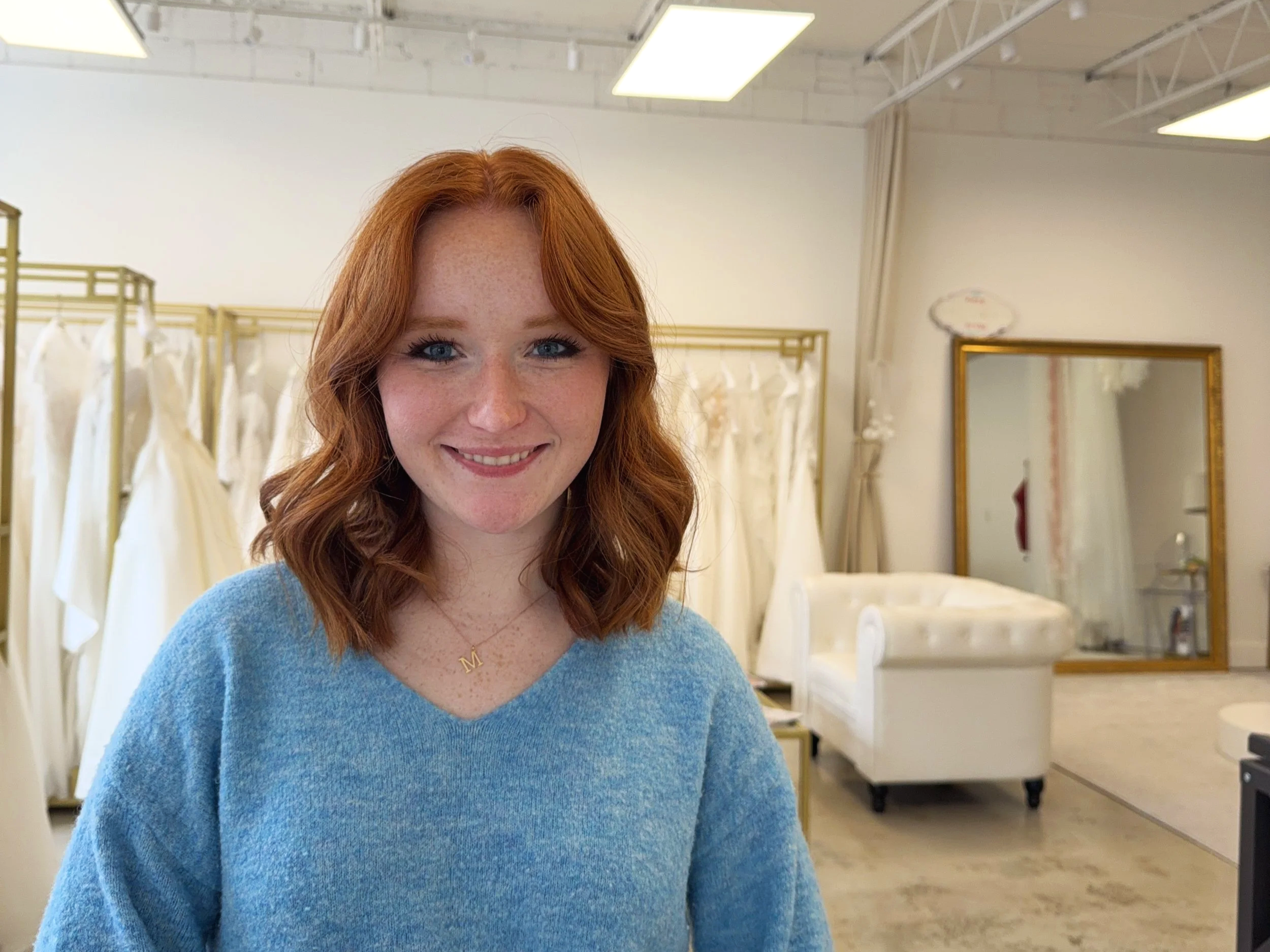 A woman with red hair, blue eyes, and freckles, smiling, wearing a light blue sweater and a gold necklace with a letter 'M' pendant, standing inside a bridal boutique with wedding dresses hanging in the background, a large mirror, and a white tufted sofa.