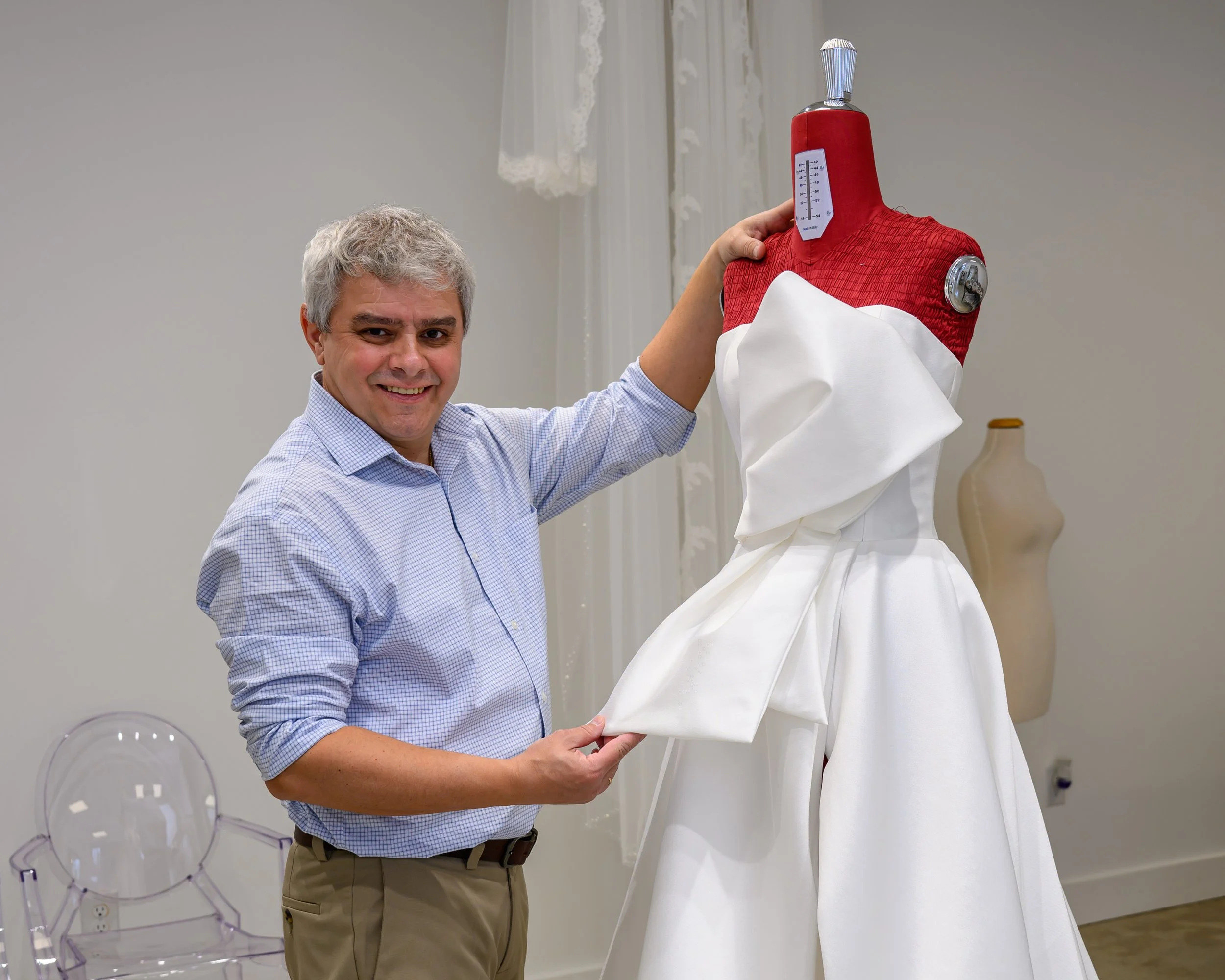 A smiling man in a light blue dress shirt and khaki pants standing next to a dress on a mannequin. The dress has a white satin skirt and a red top with a measuring tape on the neck area.