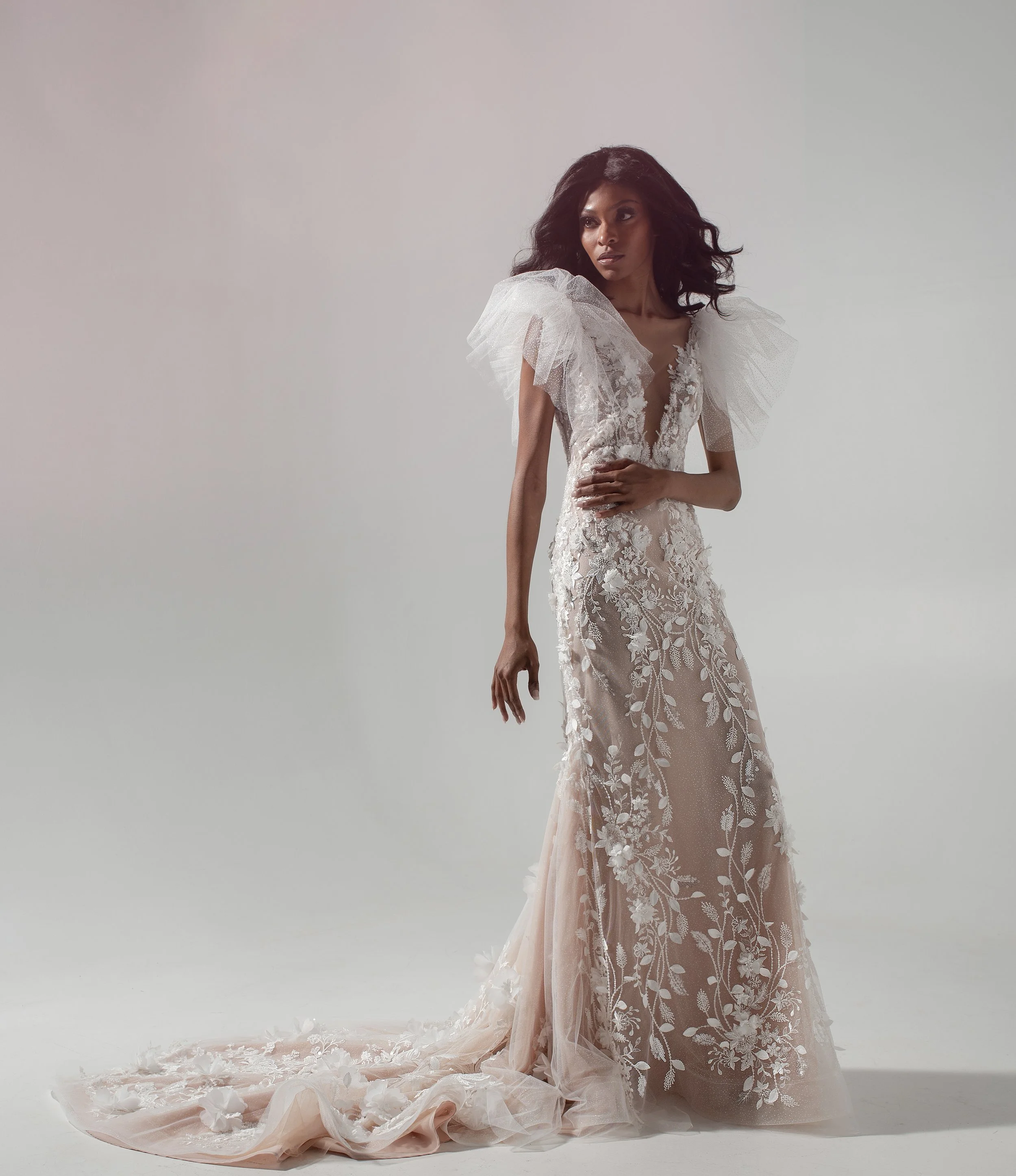 A woman in a long, white, floral embroidered gown with dramatic tulle shoulder accents, standing on a plain, light background.