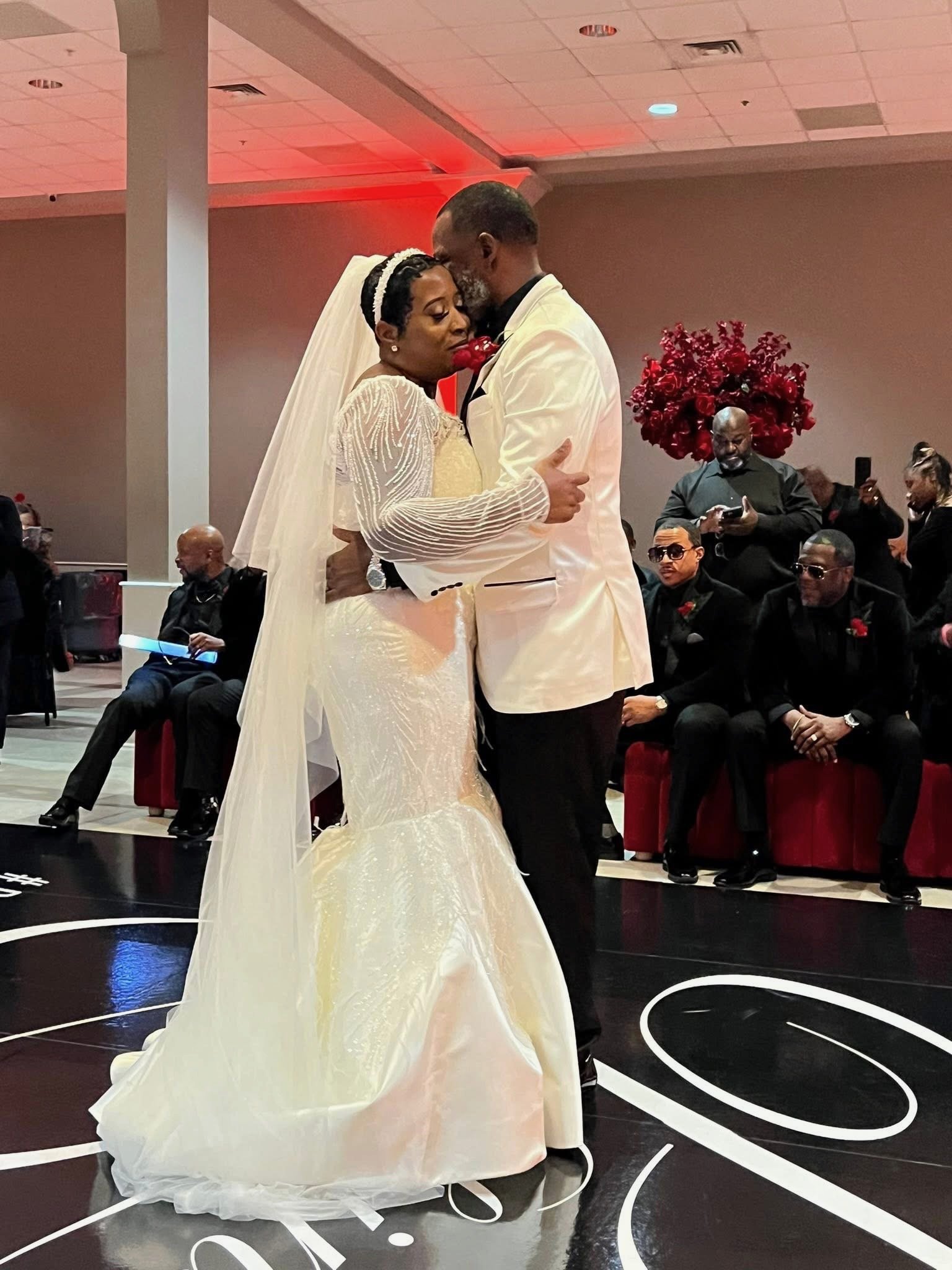 A bride and groom are dancing closely at their wedding, with the bride wearing a white dress and a veil, and the groom in a white tuxedo jacket. They are surrounded by seated guests in formal attire and an elegant venue with red and black decor.