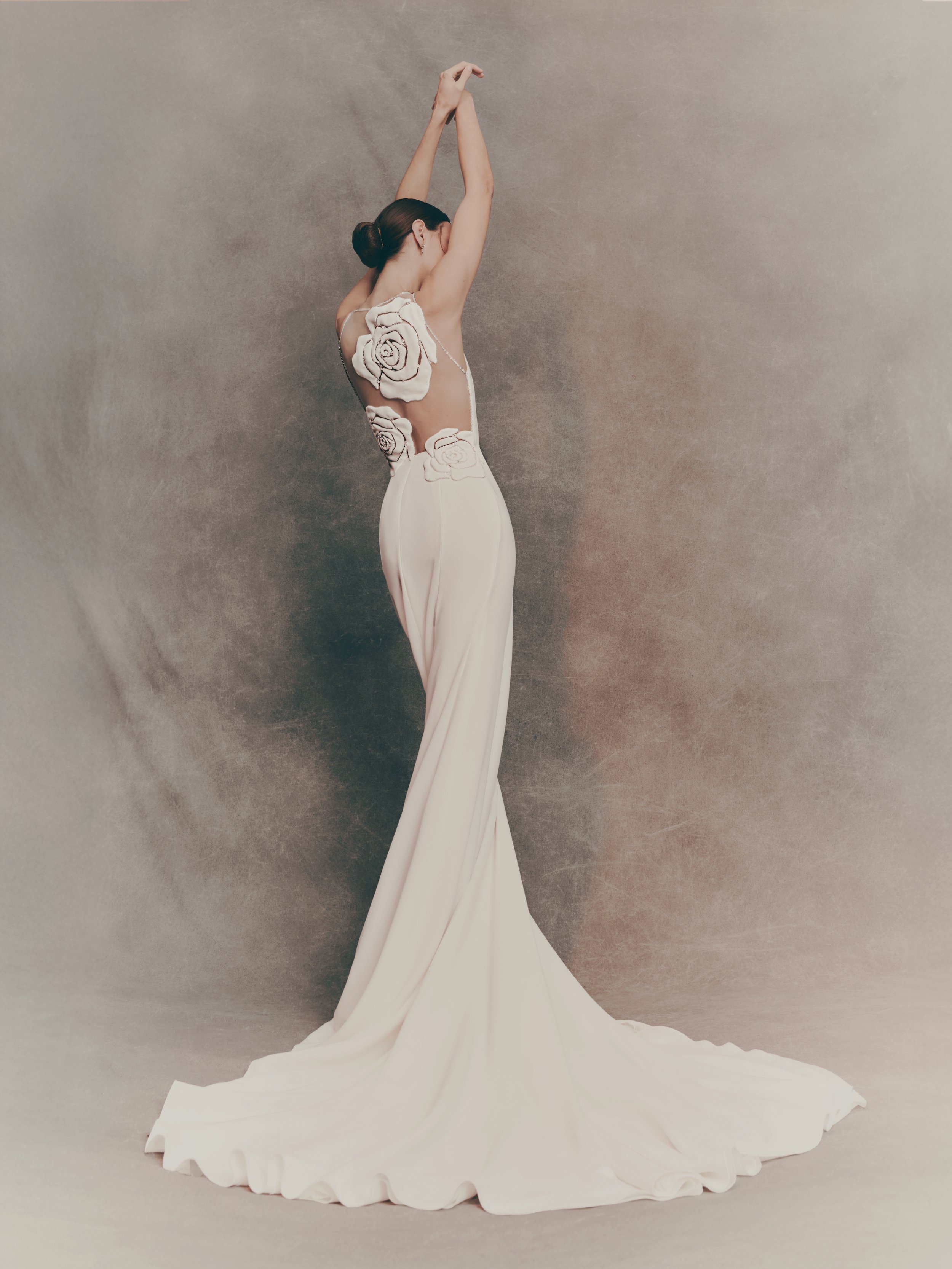 A woman in a white dress with floral lace details sits against a neutral background, with her arms raised above her head.