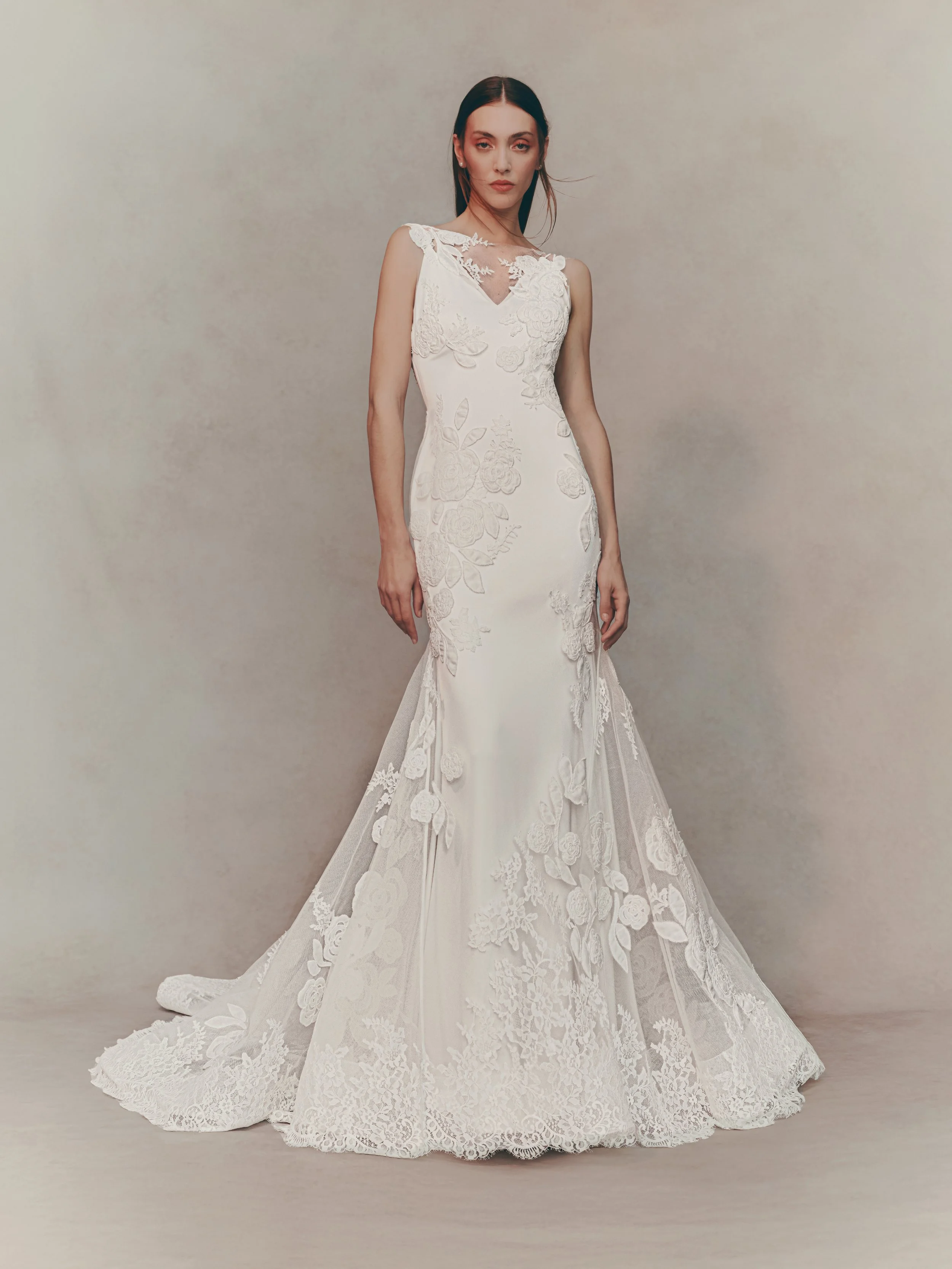 A woman wearing an elegant white lace wedding dress with floral embroidery, standing against a plain light-colored background.