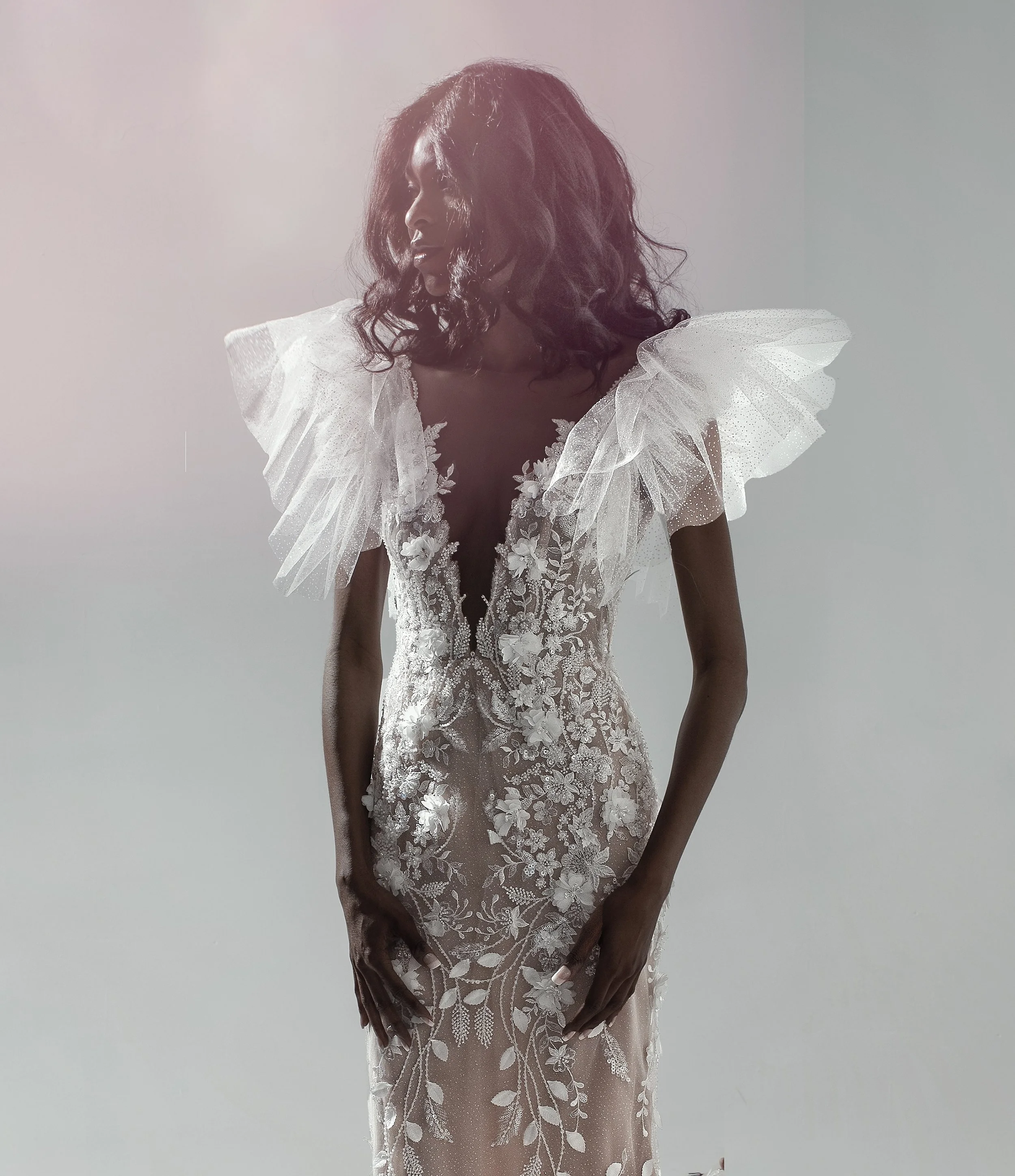 A woman wearing a white, intricately embroidered dress with ruffled shoulder details, standing against a plain background with soft lighting.