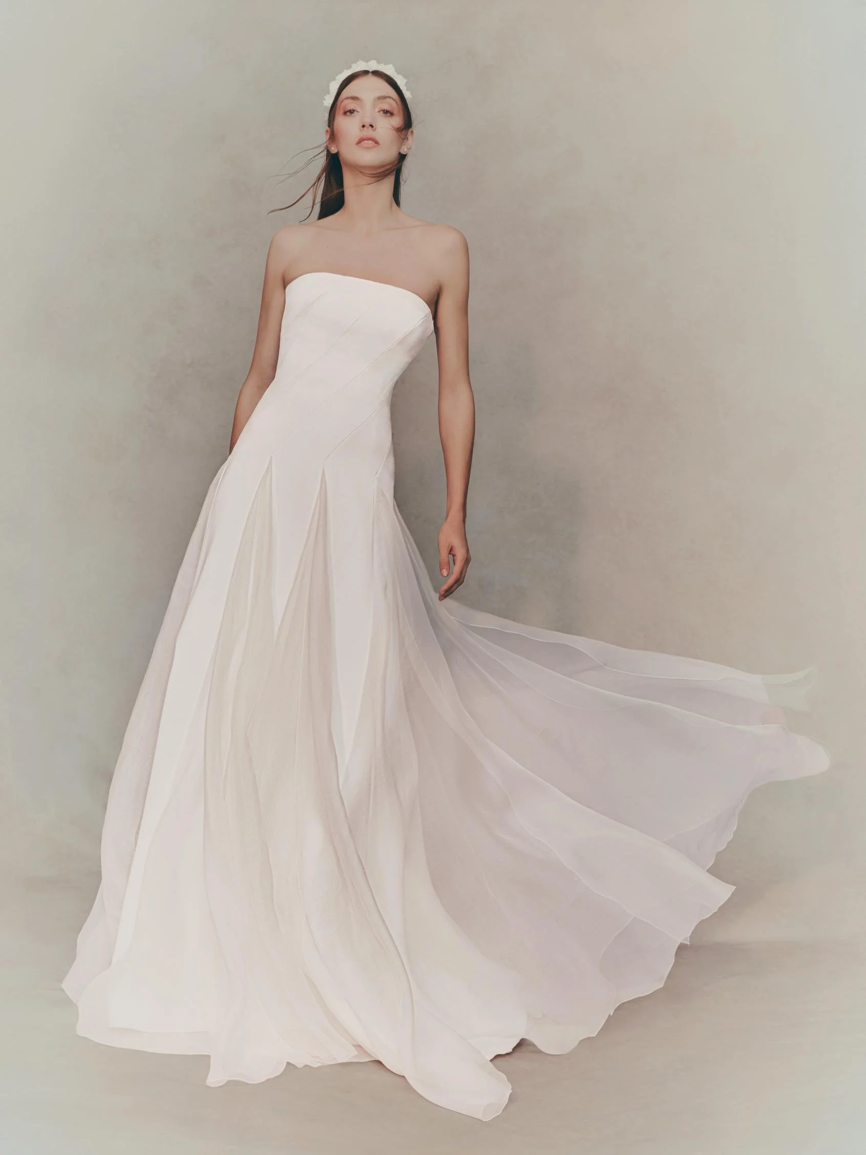 A woman in a strapless white wedding dress with a flowing skirt, standing against a neutral background.