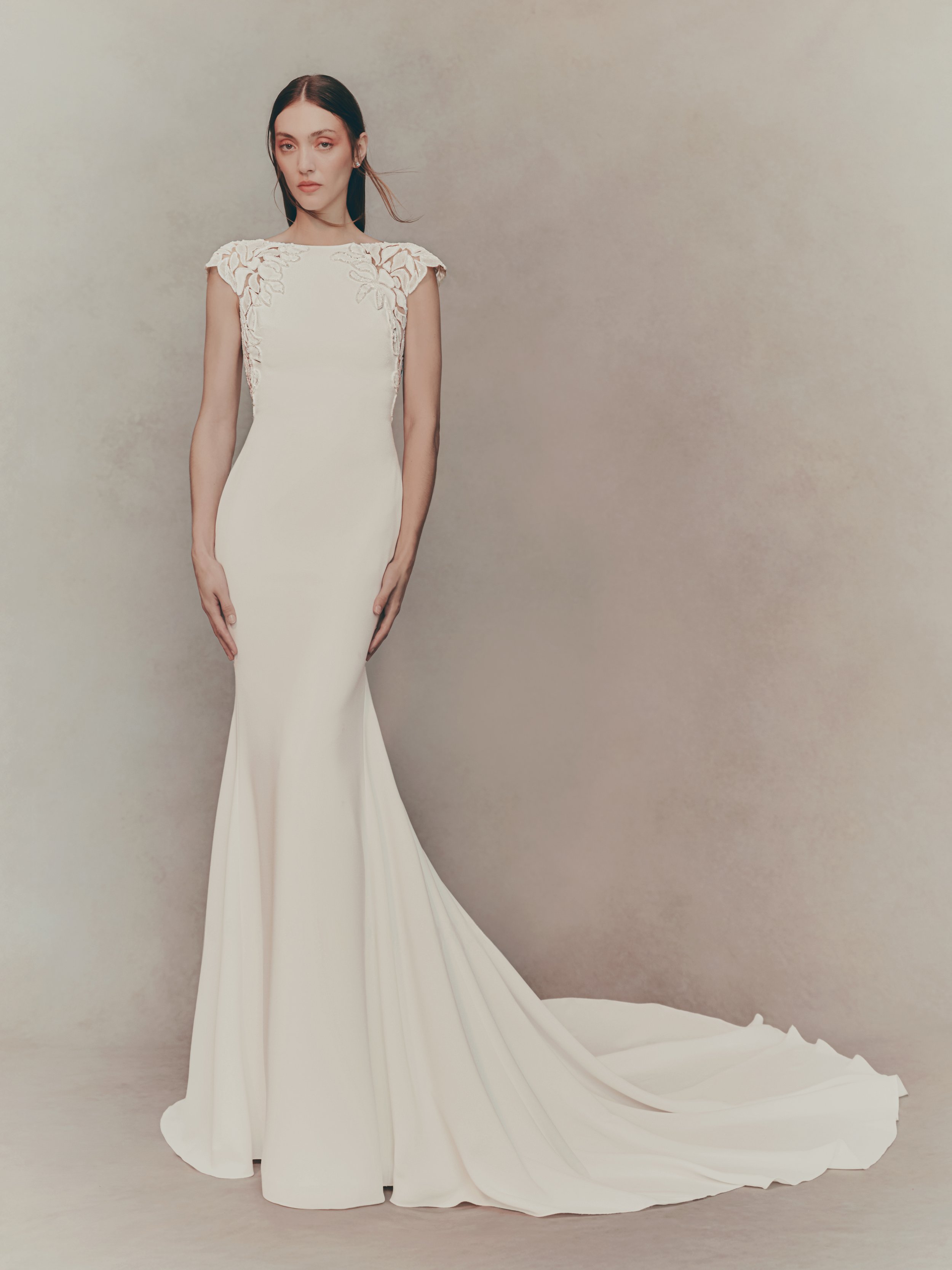 A woman in a white wedding gown with lace details on the shoulders, standing against a neutral background.