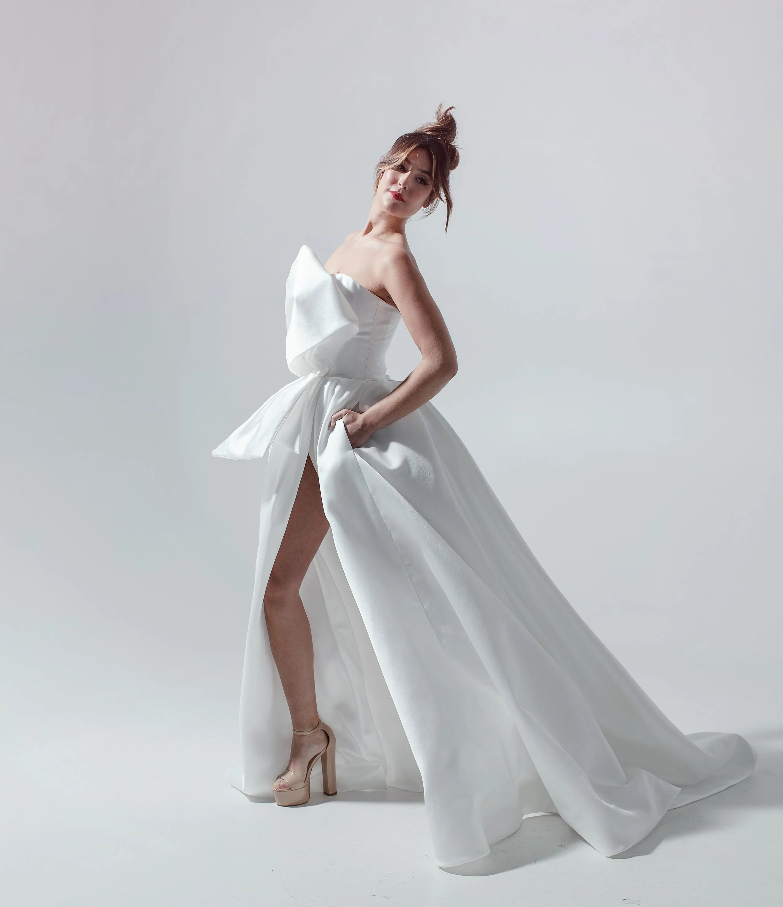 A woman wearing a white strapless gown with a high slit, high heels, and with her hair styled in a high bun, standing against a plain light gray background.