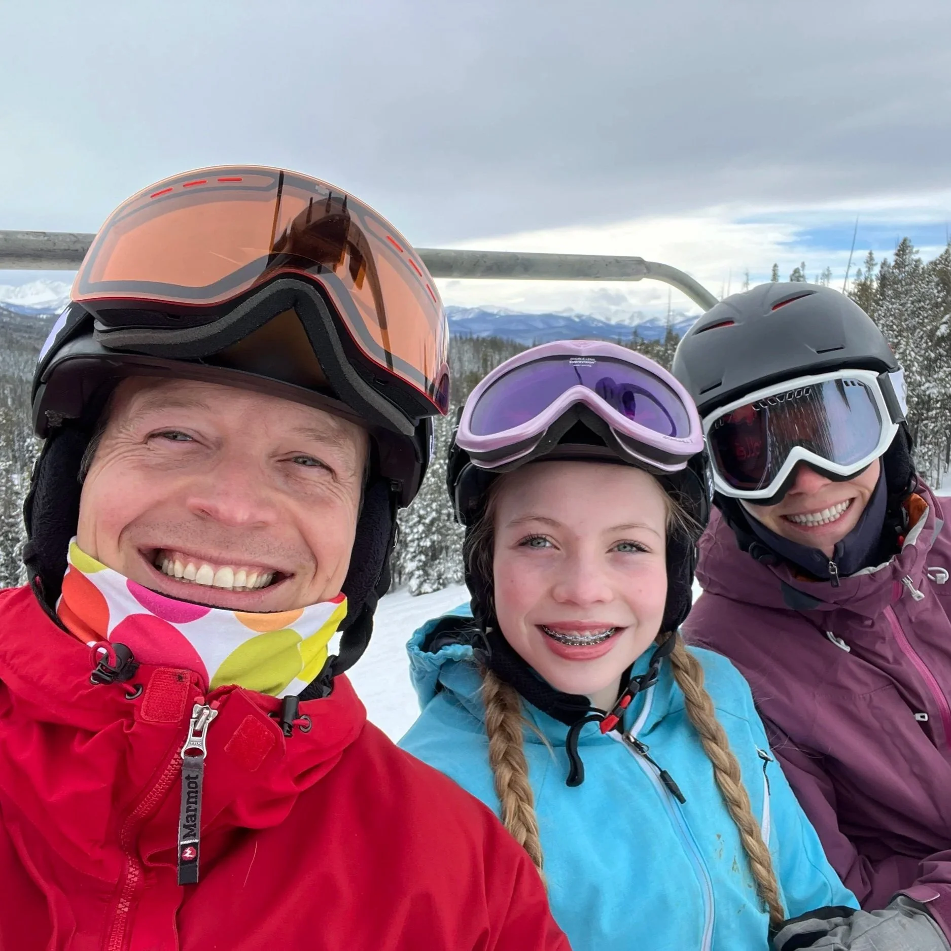 Three people smiling in winter gear and ski helmets on a snowy mountain, with ski lifts and trees in the background.