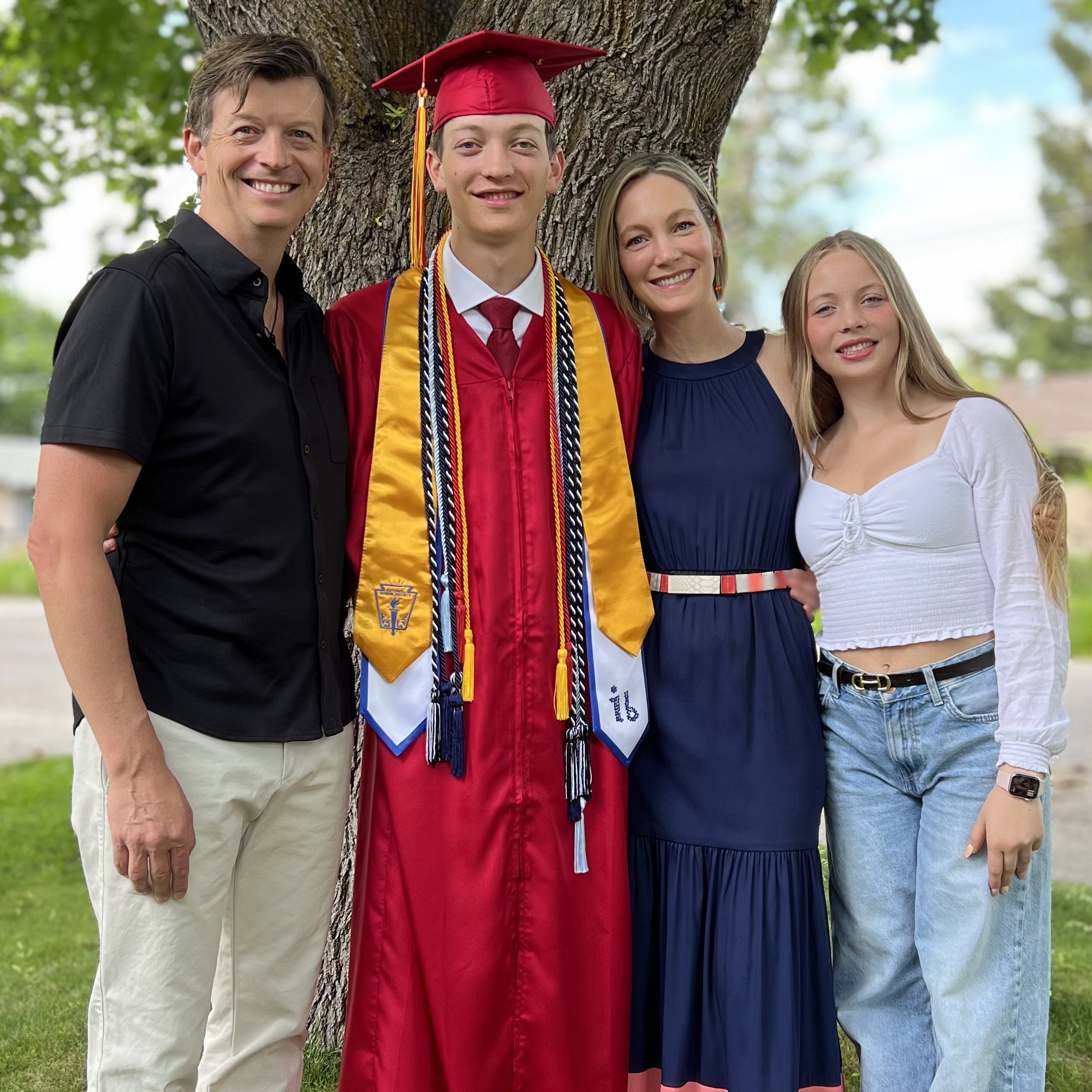 A family celebrating a graduation outdoors, with a boy in a red cap and gown, surrounded by two women and a man, all smiling.