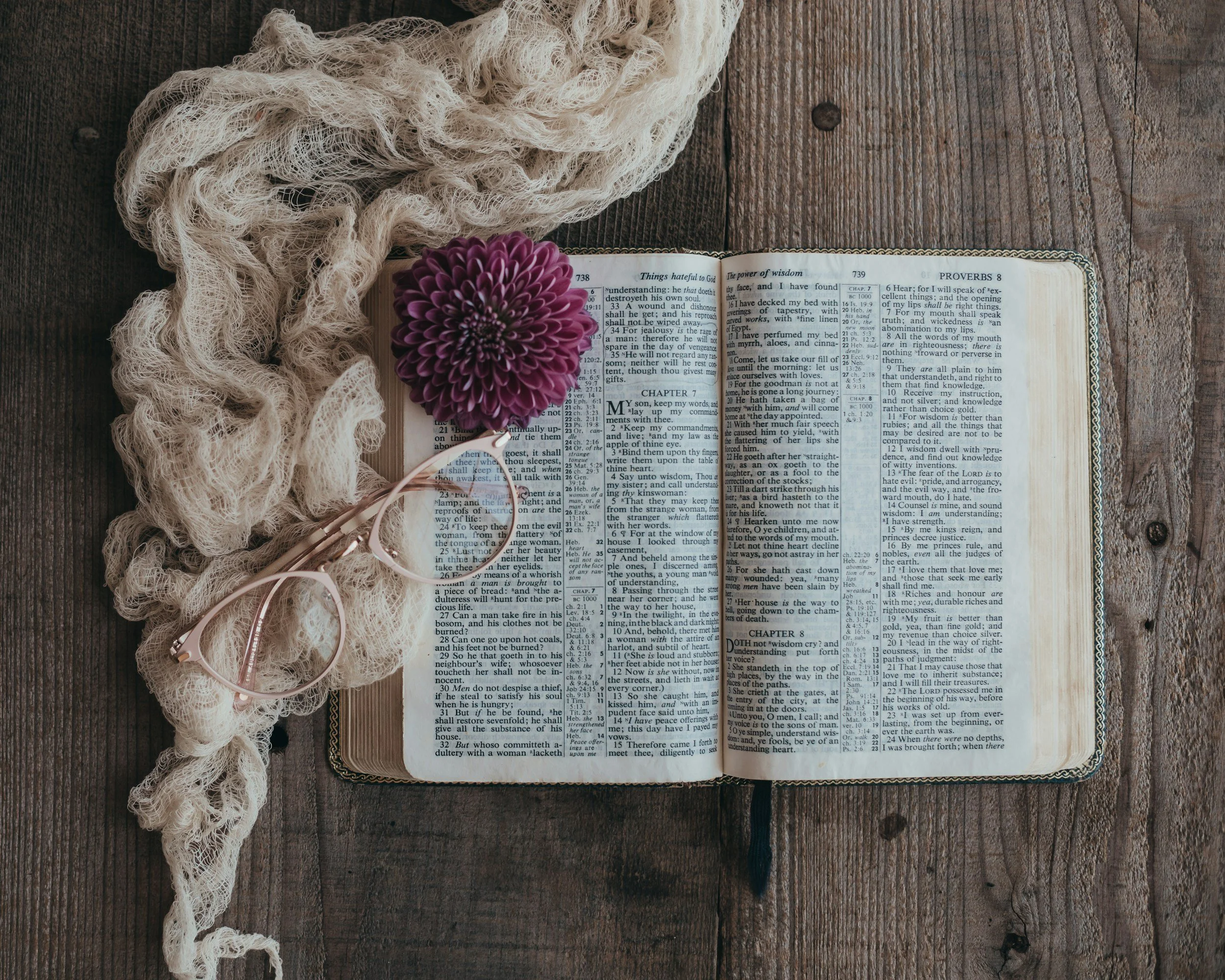 An open Bible resting on a wooden surface with a purple flower on top. A beige lace scarf and a pair of pink reading glasses are also placed on the surface.