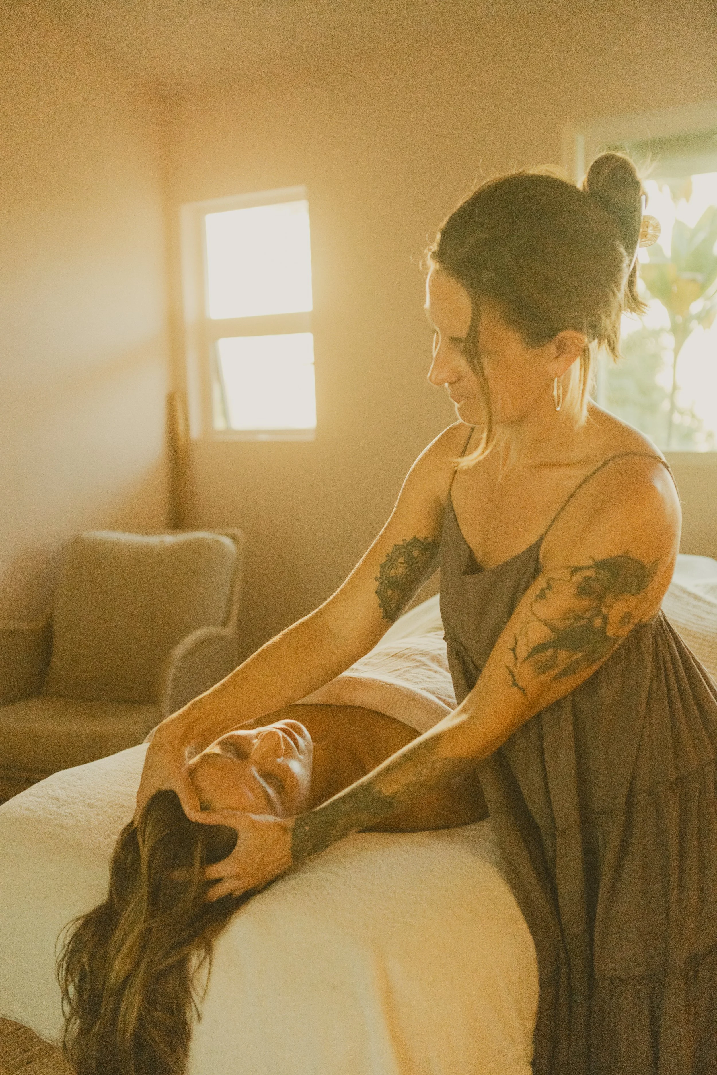 A woman giving a massage to another woman on a bed in a sunlit room.
