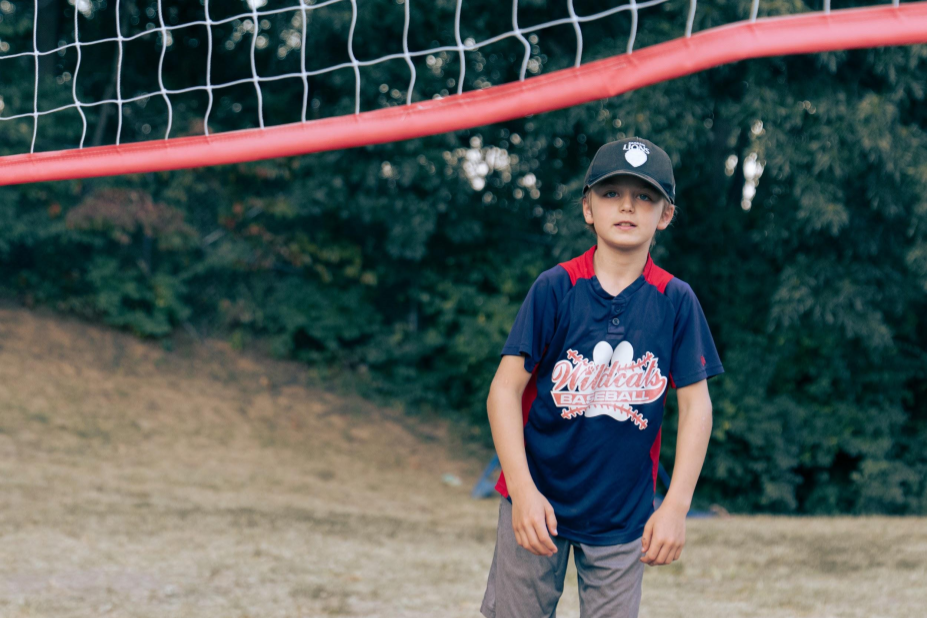 Young boy standing on a grassy field near a volleyball net, wearing a black cap and a navy blue and red Wildcat baseball jersey.