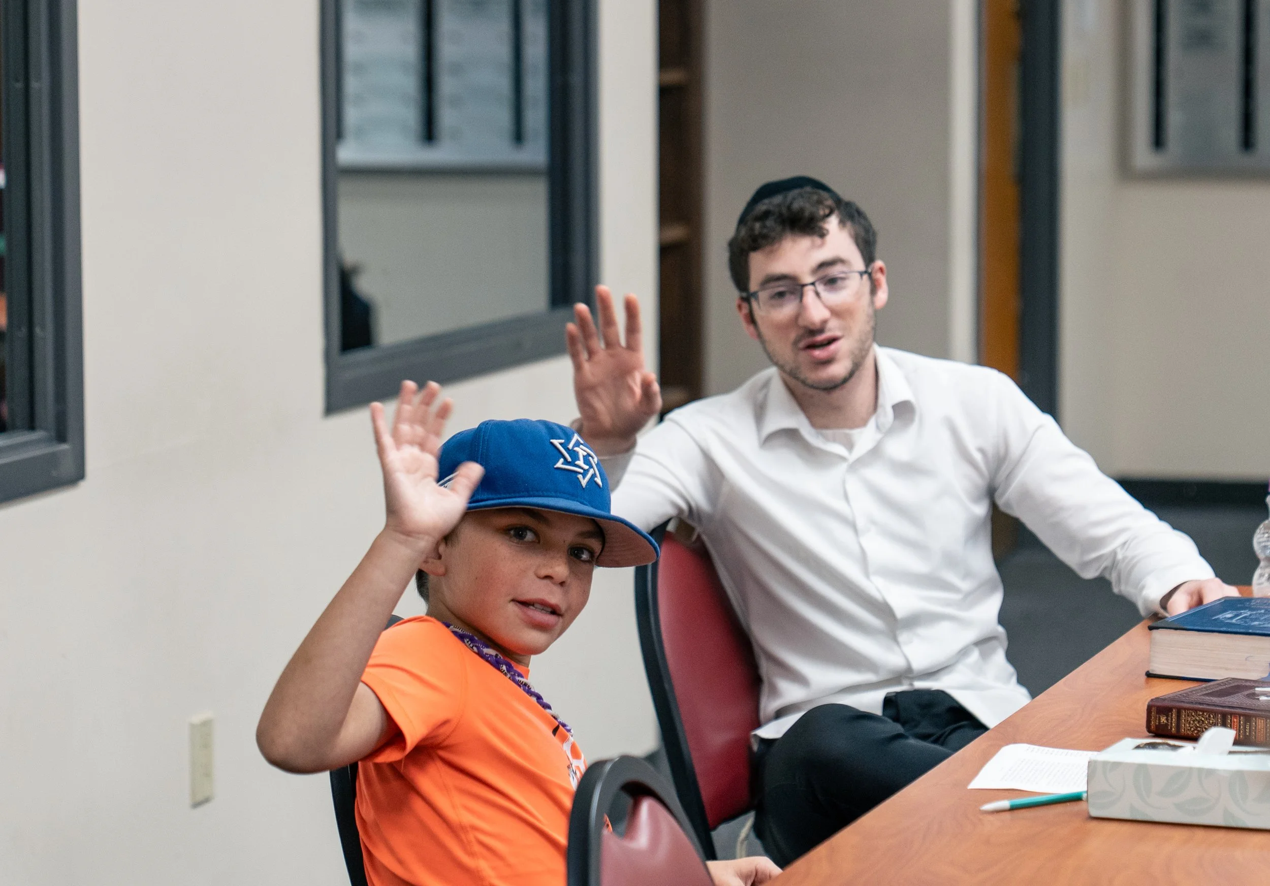 A young boy in an orange shirt and blue baseball cap waving, seated next to a man in a white shirt and black yarmulke, both seated at a wooden table in a room with windows and books.