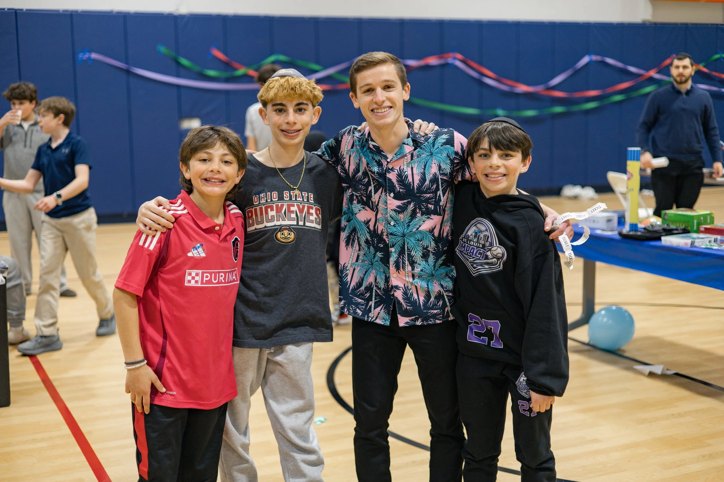 Four young boys standing together in a gym, smiling, with a blue wall and colorful streamers in the background.