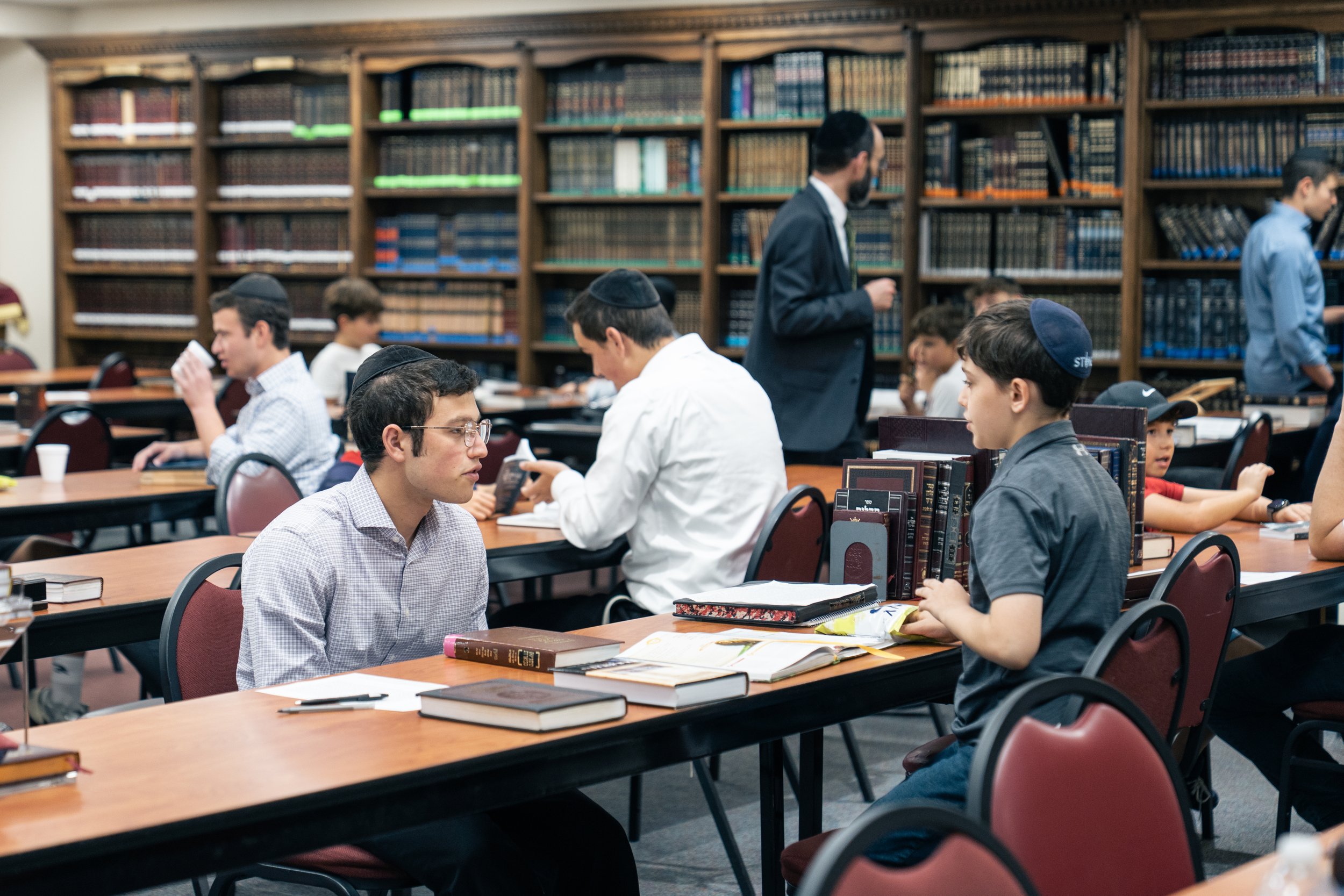 Young boys in traditional Jewish attire studying and using electronic devices in a library with bookshelves filled with religious texts.